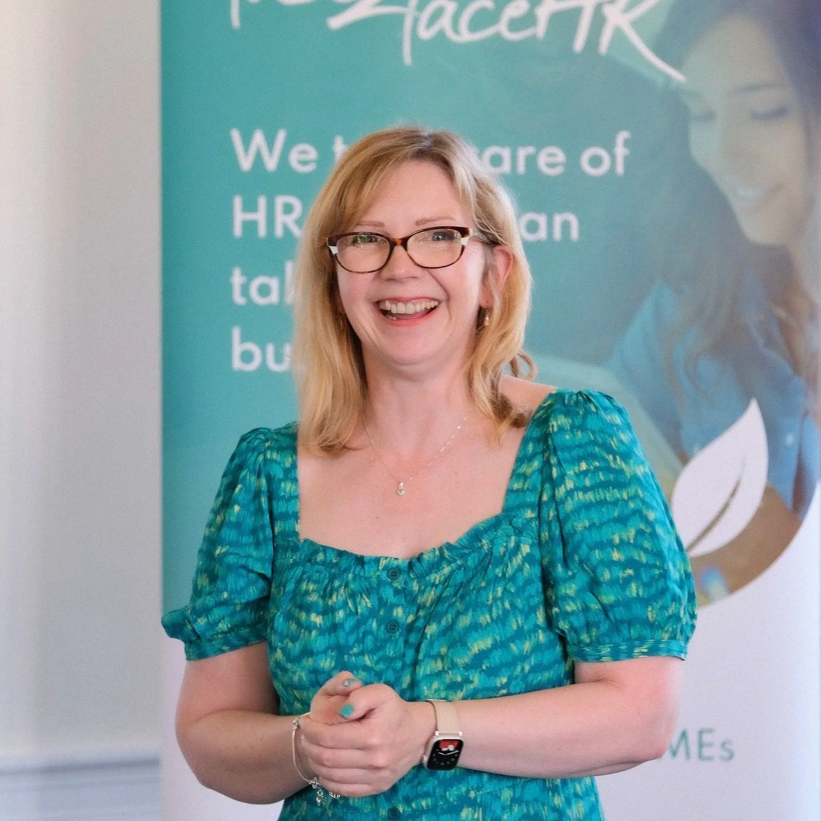 Eleanor Deem smiling wearing a teal patterned dress, standing in front of a face2faceHR banner.