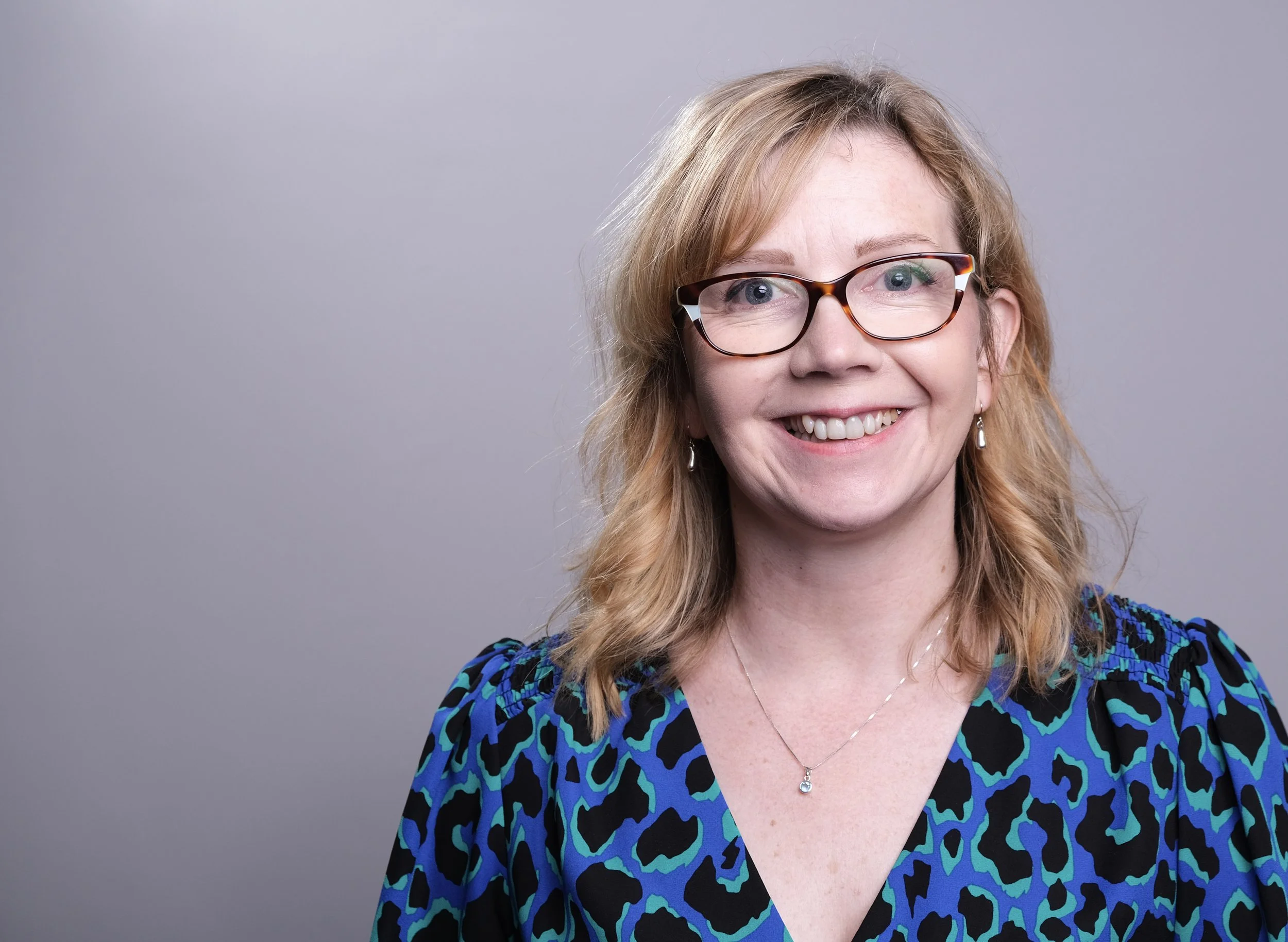 Portrait of Eleanor Deem, wearing a blue and black patterned top, against a plain grey background.