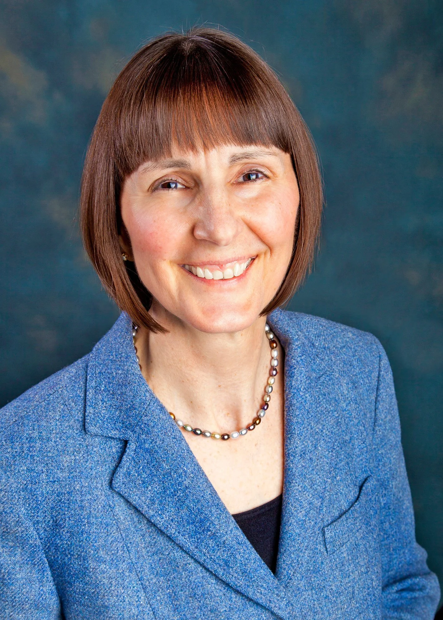 Close-up of a smiling woman with short brown hair, wearing a blue blazer and a multicolored pearl necklace, against a dark, blurred background.
