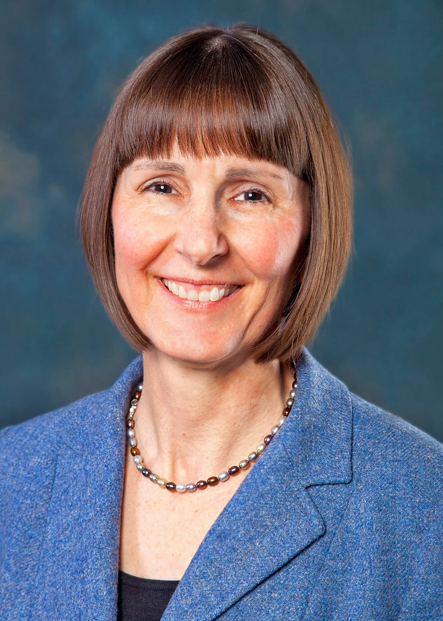 Close-up portrait of a smiling woman with short brown hair, wearing a blue blazer and pearl necklace, against a dark background.