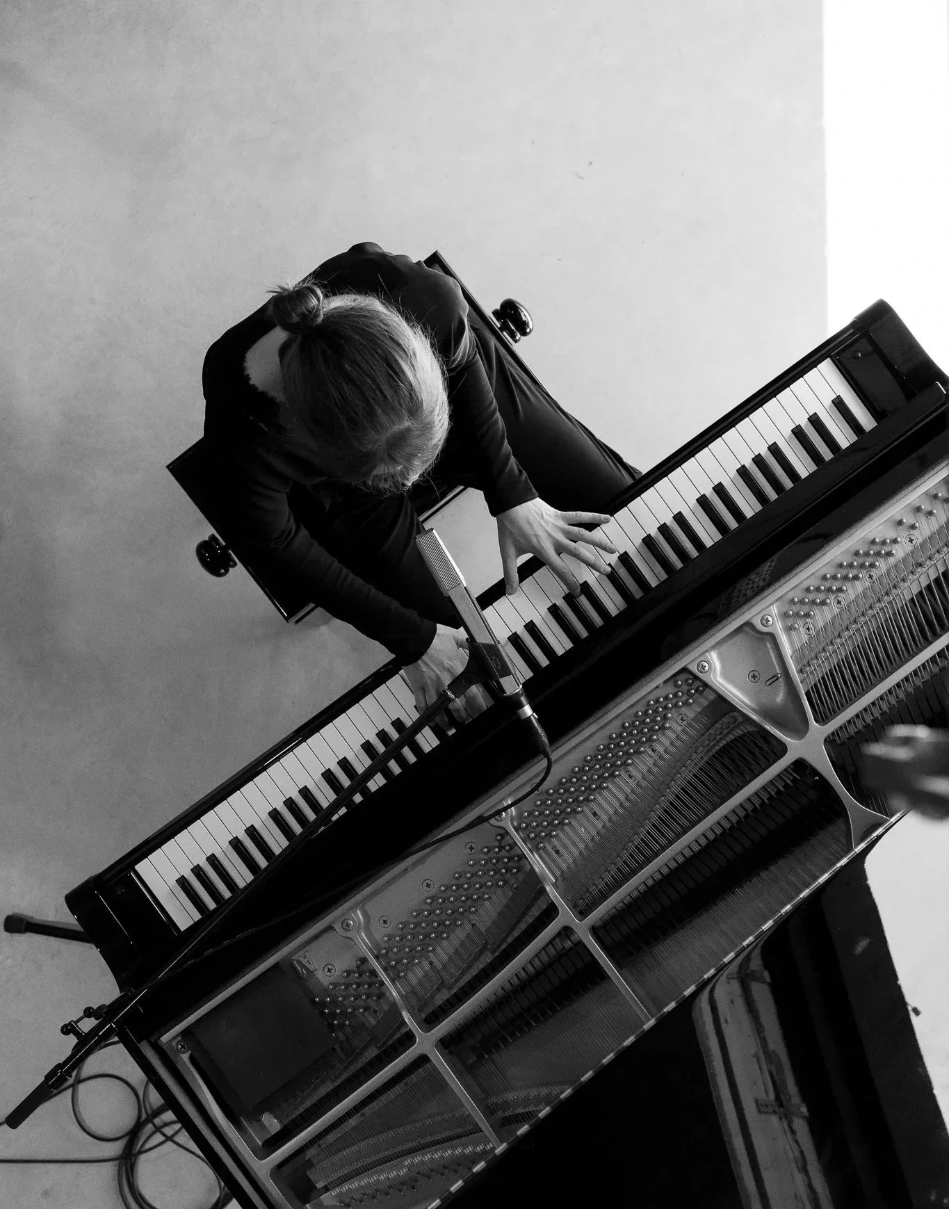 A person with long hair in a bun playing a grand piano, viewed from above in black and white