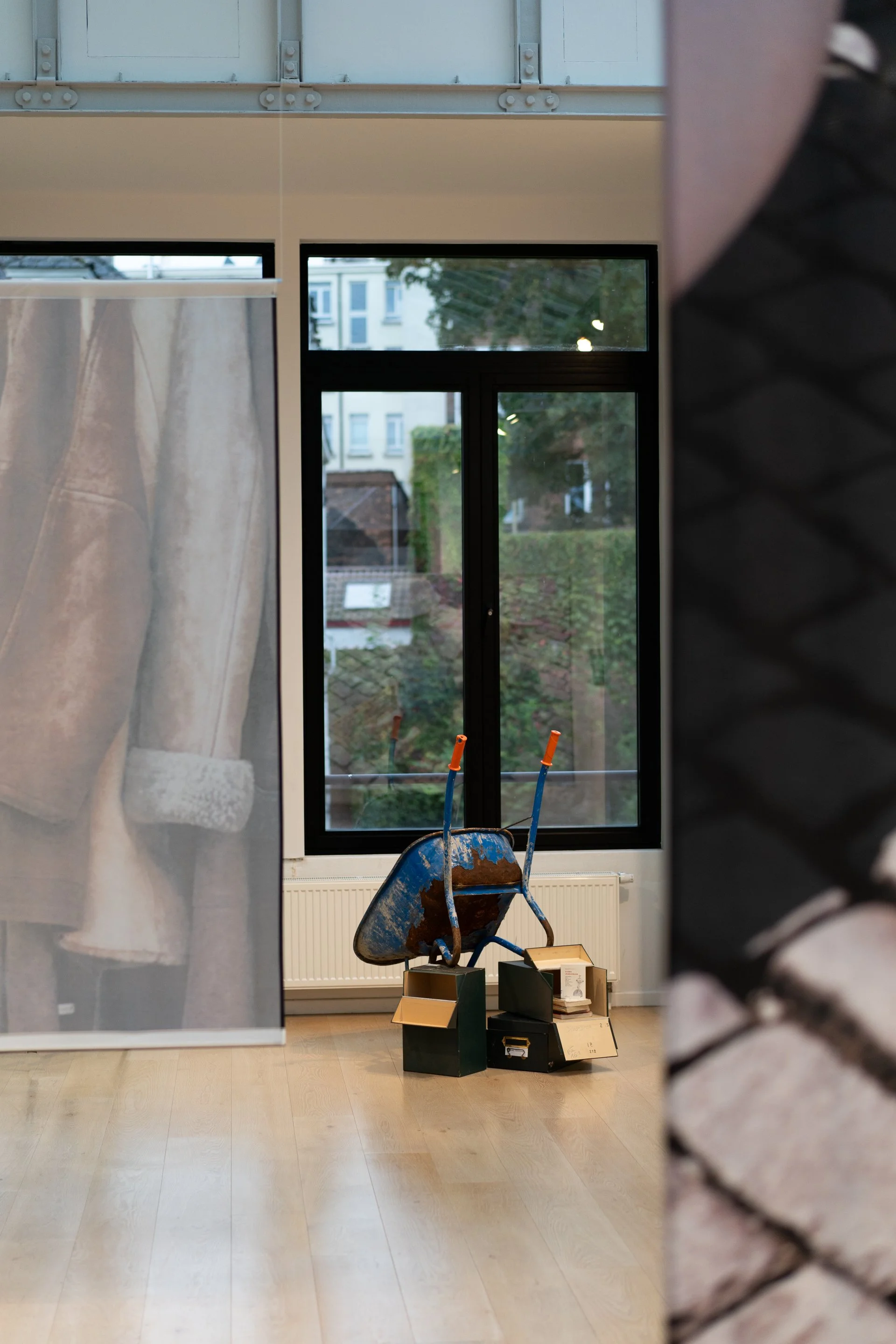 Indoor room with large window, blue wheelbarrow filled with dirt, and boxes on the floor. Part of a curtain on the left and a brick wall on the right are visible.