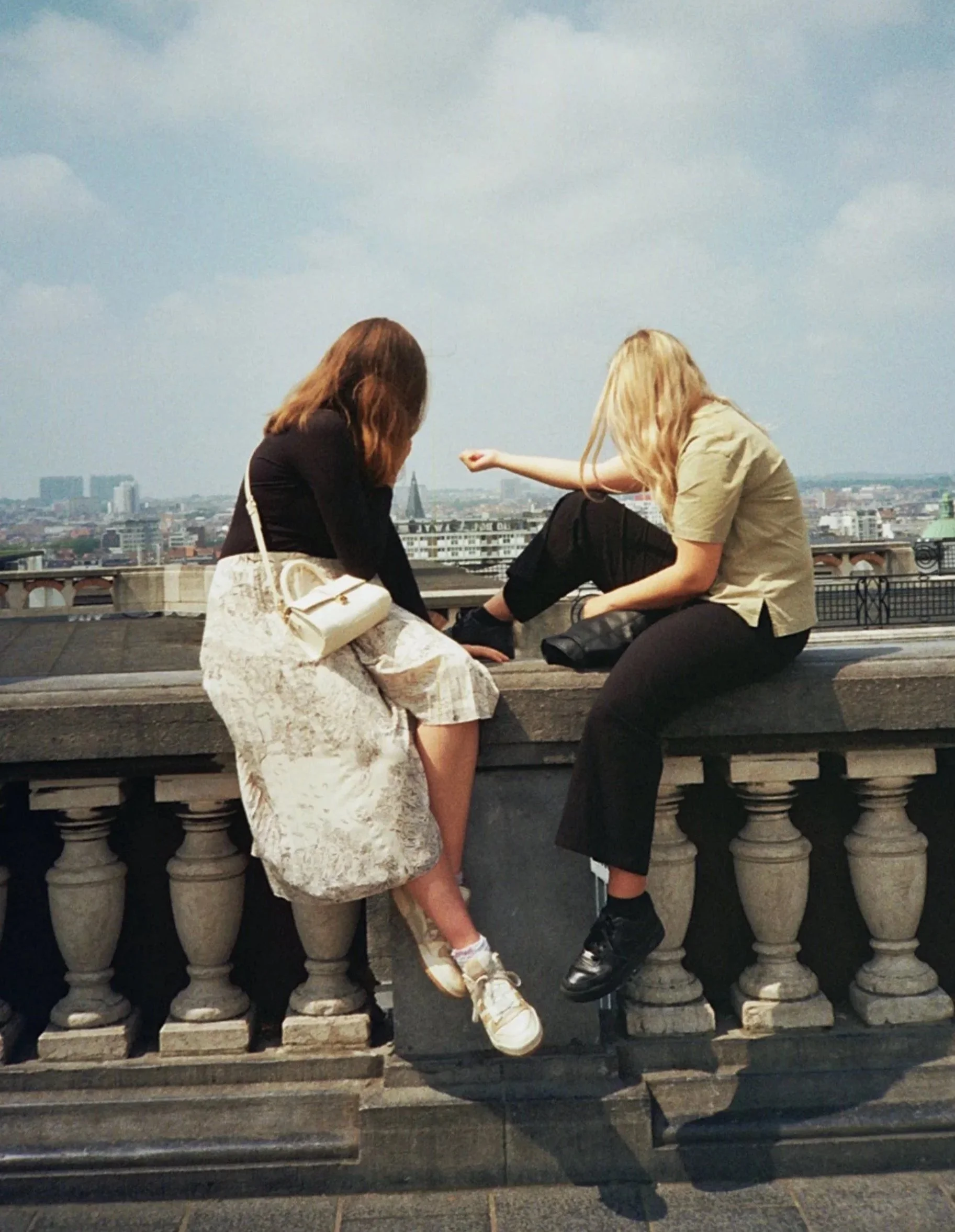 Two women sitting on a ledge of a balcony overlooking a city skyline in Paris, with some clouds in the sky.