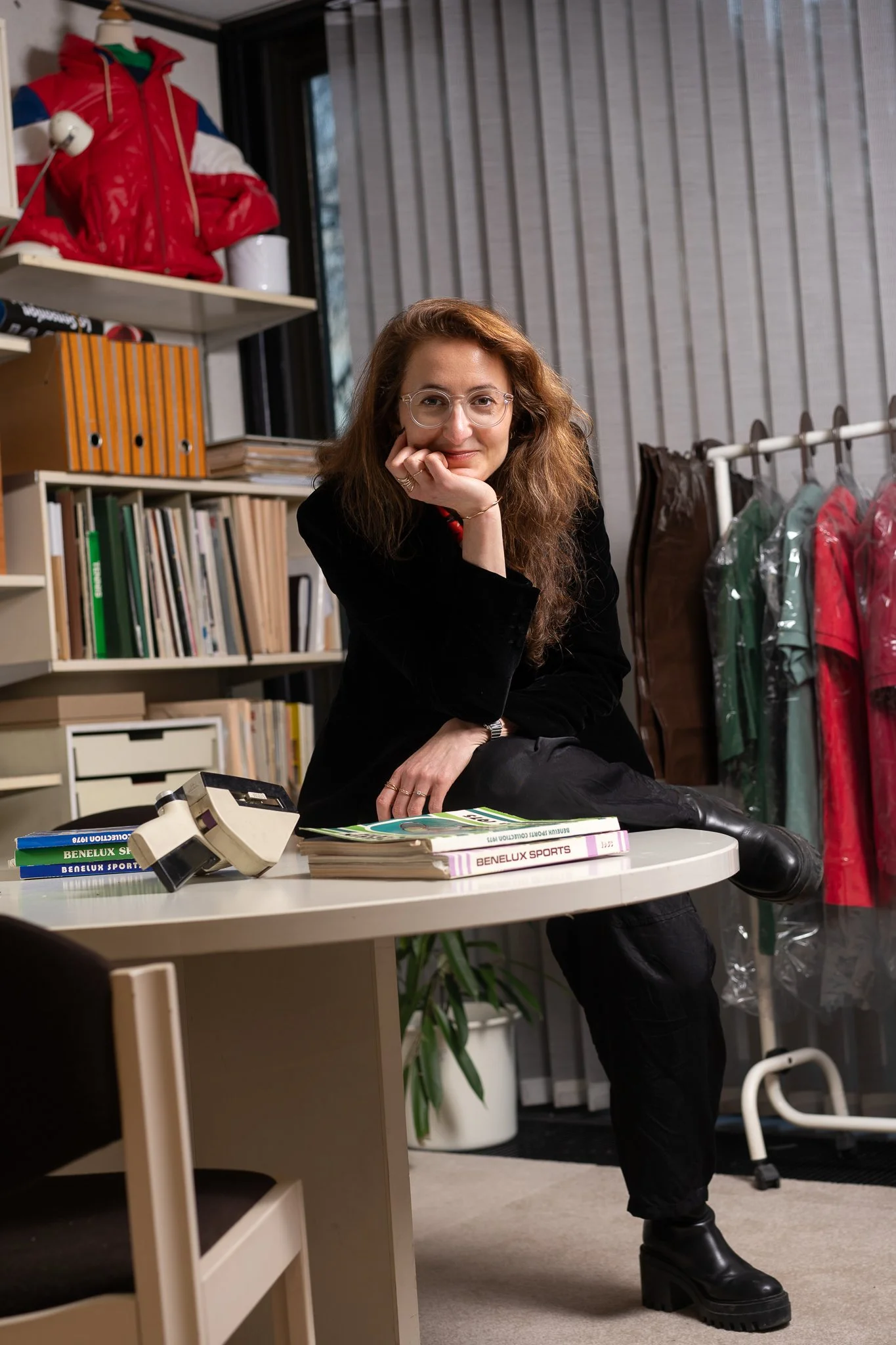 A woman with long, wavy red hair and glasses, smiling and resting her face on her hand, seated at a desk with books and a vintage camera. Behind her are shelves filled with binders and books, and clothing hanging on a rack, in a room with gray vertical paneling and a window.