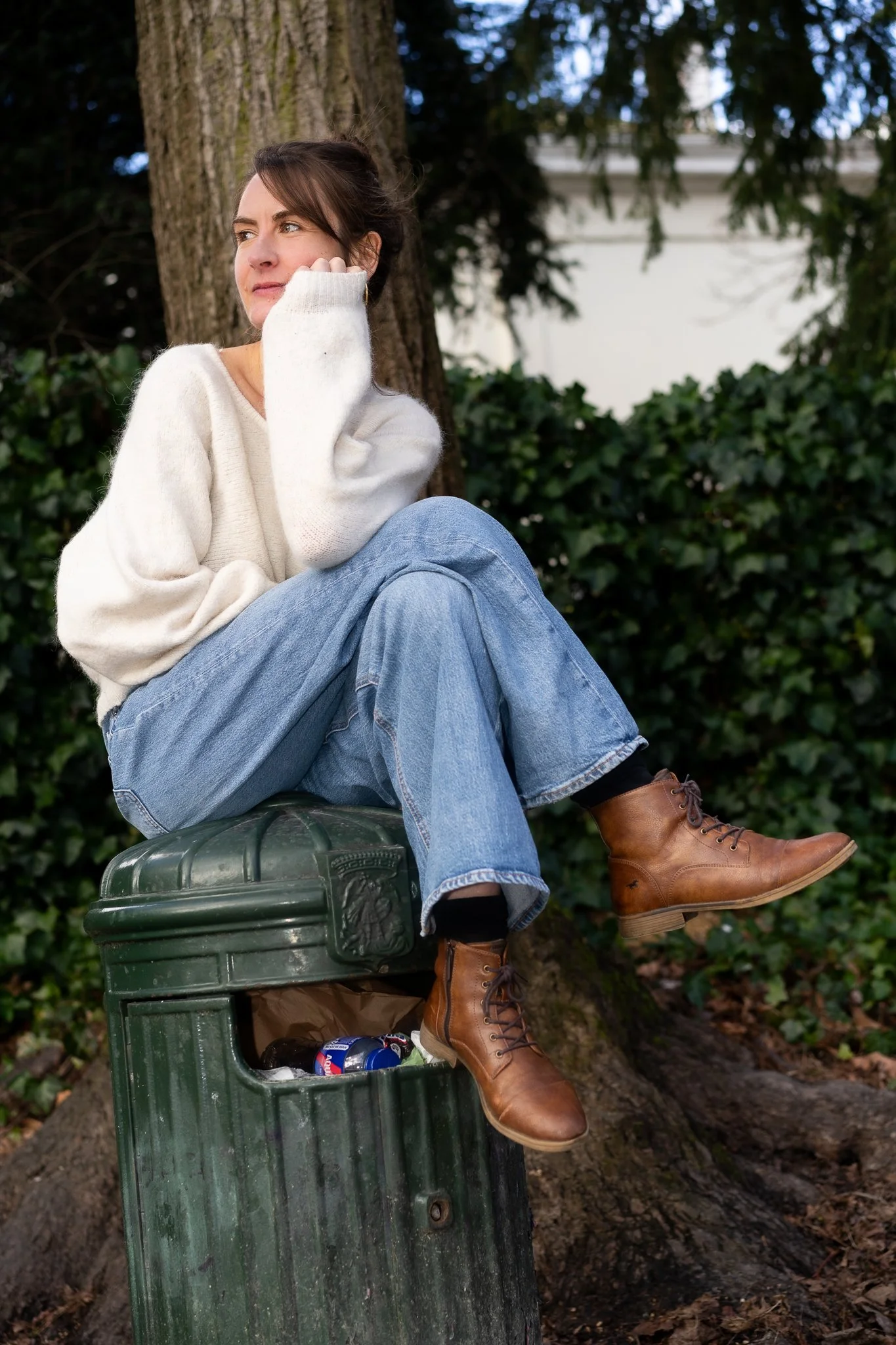 A woman sitting on a green trash can next to a tree in a park, with one leg crossed over the other. She is wearing a white sweater, blue jeans, and brown boots, with her hand resting on her cheek and looking away thoughtfully.