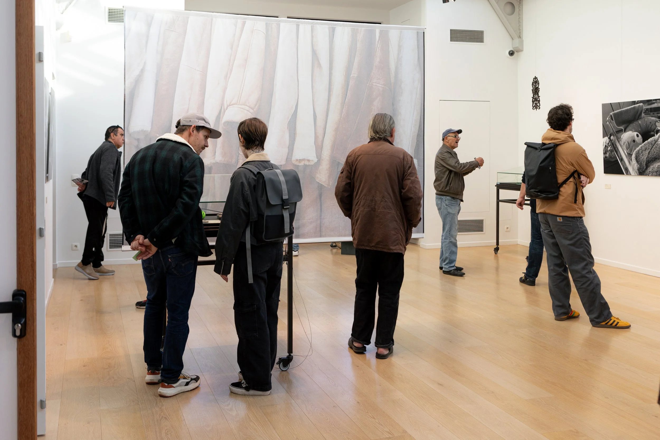 People standing in line in an art gallery, looking at artwork on the walls and a large painting or photograph in the background.