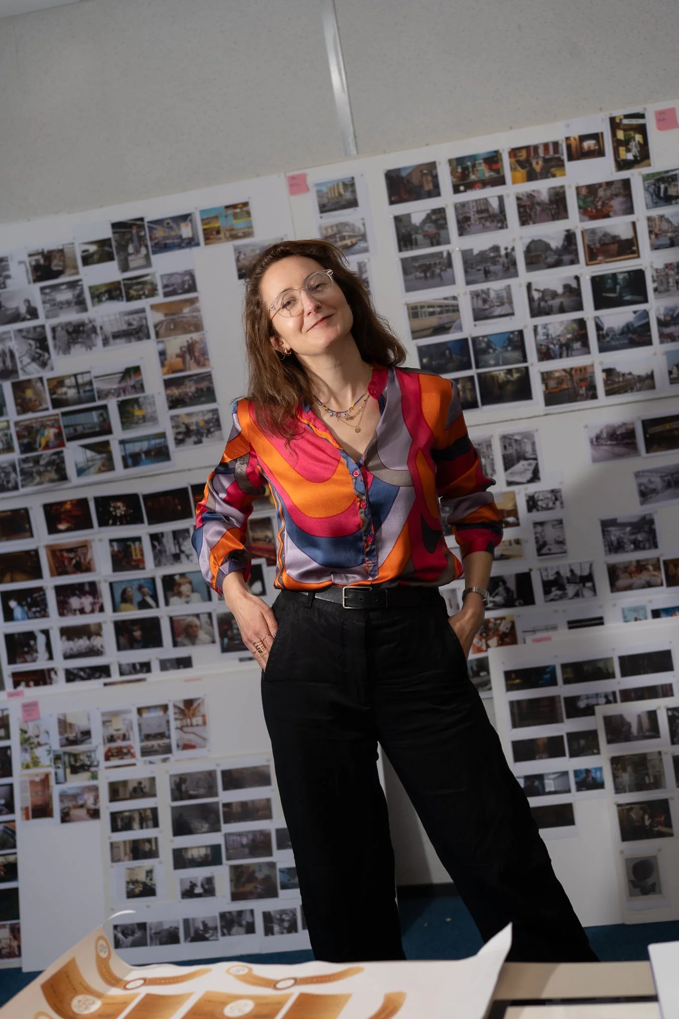 A woman with glasses and long hair, wearing a colorful patterned shirt, standing in front of a wall covered with many small photographs or images.