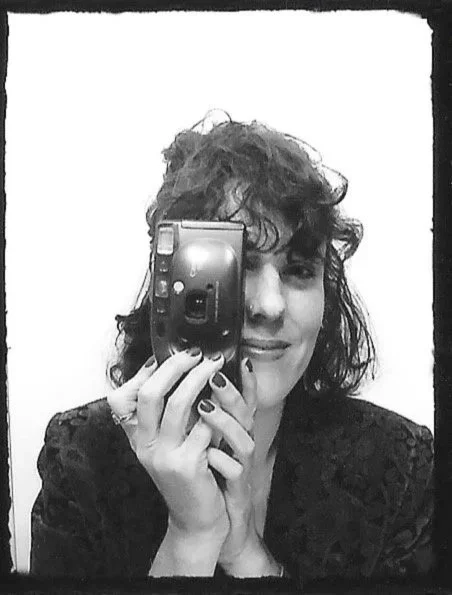 A woman with curly hair is taking a selfie in a mirror with a camera, smiling slightly.