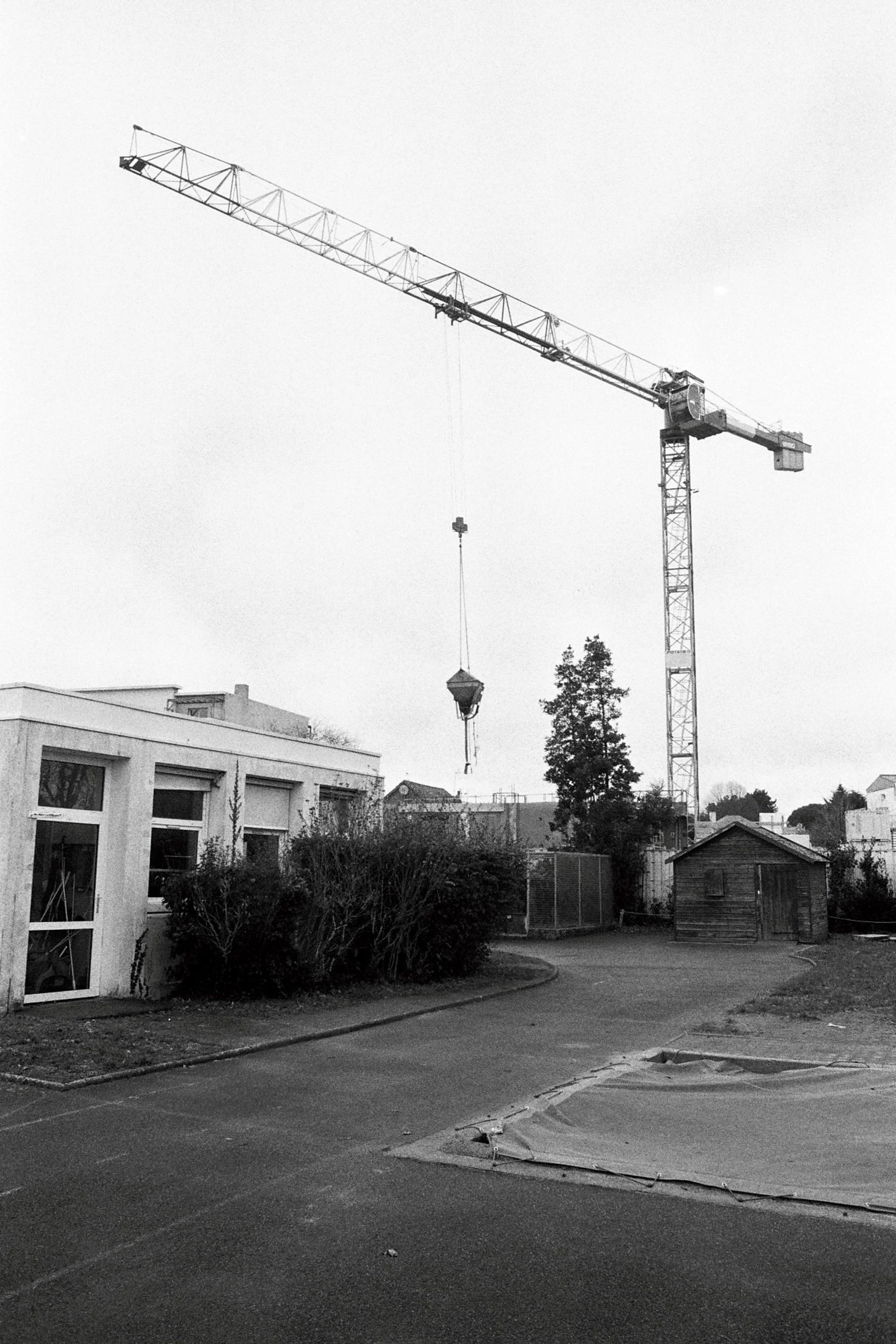 A black and white photo of a construction site with a tall crane lifting materials. There are buildings, trees, and a partially paved street in the foreground.