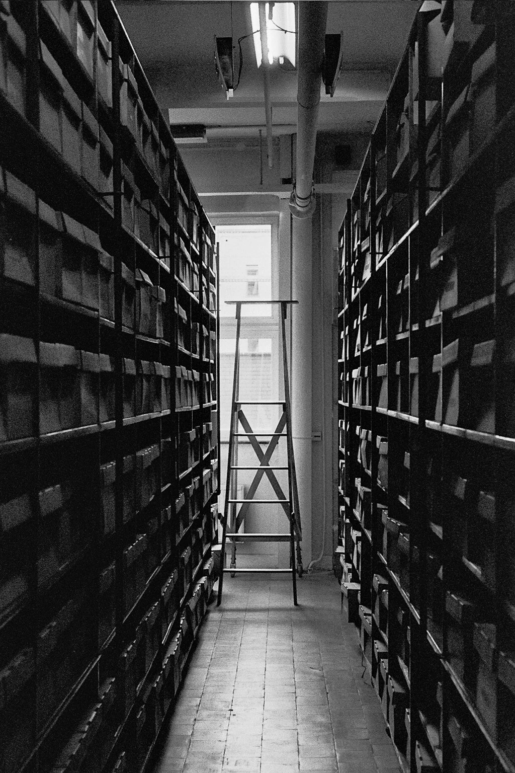 Black and white photo of a narrow aisle between tall shelves filled with files or binders, with a ladder in the background near a window.