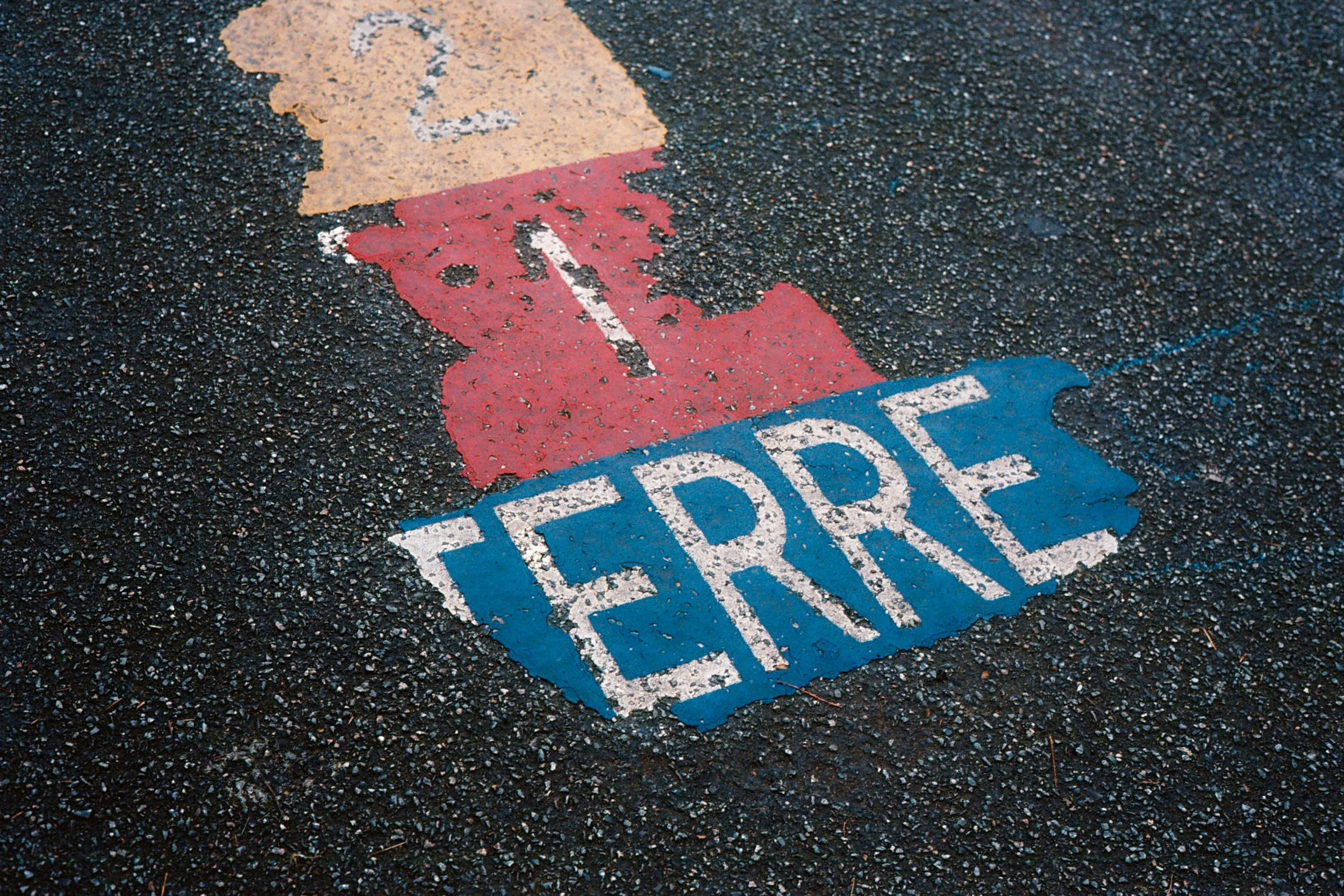 Faded and peeling painted parking space marking on asphalt that says 'TICKET' with a red arrow pointing upward and blue arrows to the sides.
