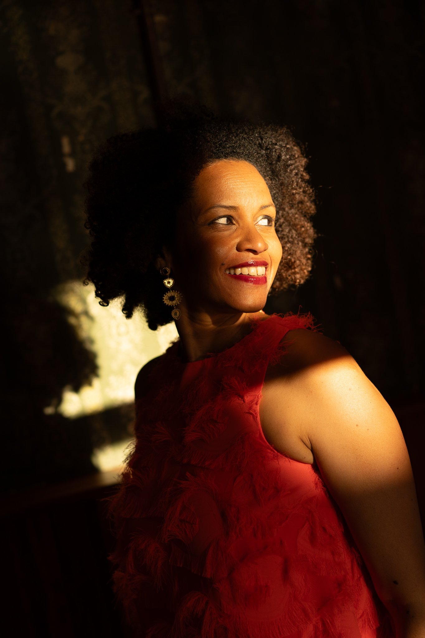 A woman with curly hair and earrings smiling and looking to the side, wearing a red textured dress in dim lighting.