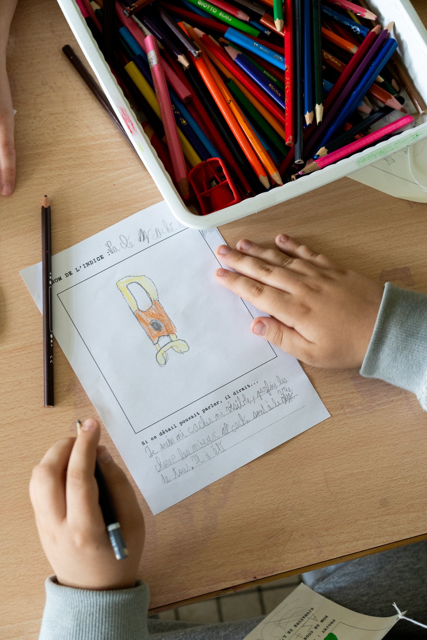 Children's hands working on a drawing and writing activity at a desk, with a box of colored pencils and a sheet of paper showing a hand-drawn image.