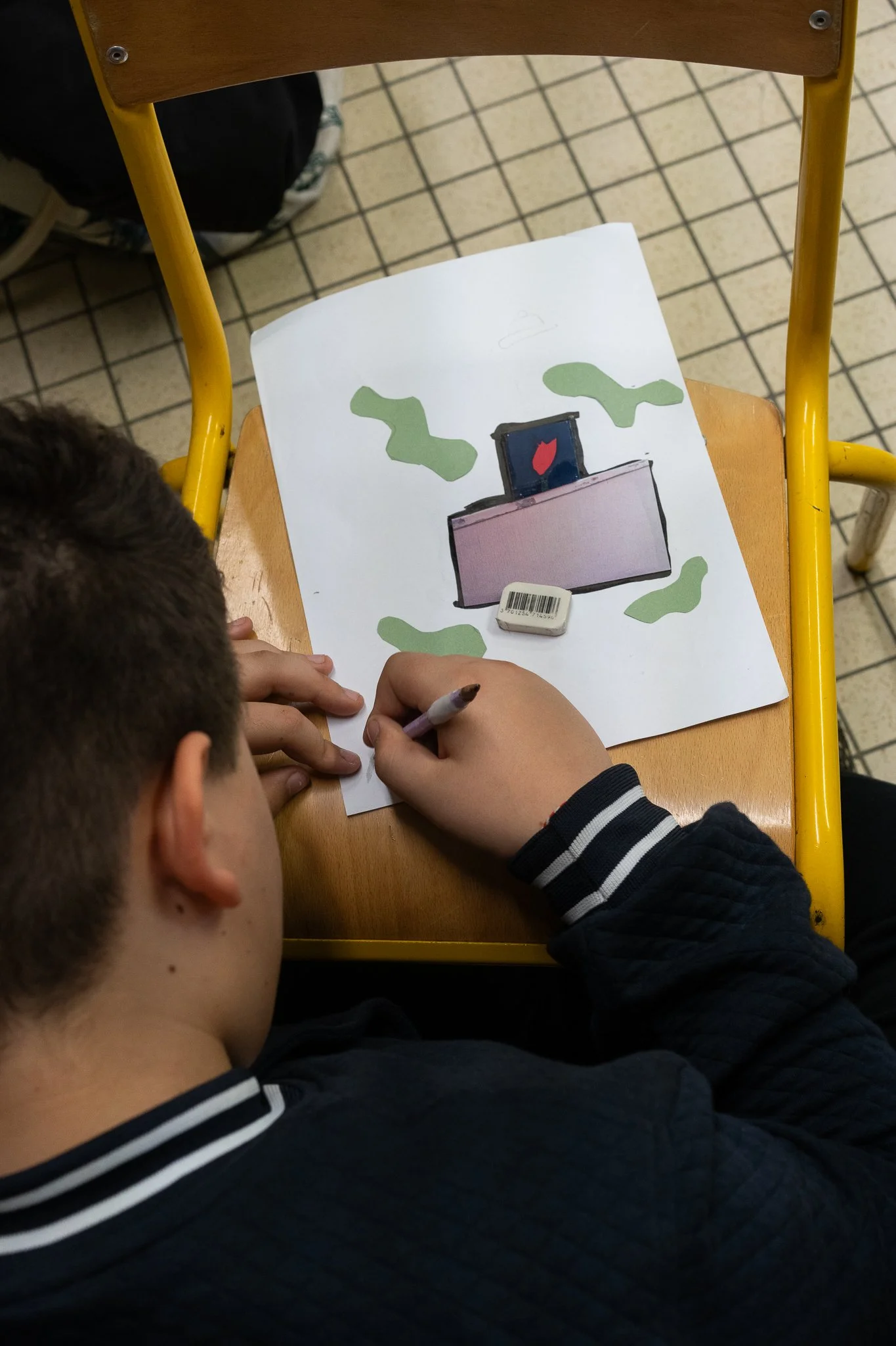 A child with short dark hair and a black and white striped sweater sitting at a wooden school desk. The child is drawing or coloring on a sheet of paper that has a picture of a spray bottle and some green shapes. The floor is tiled with a brown and cream pattern. A backpack is partially visible in the background.