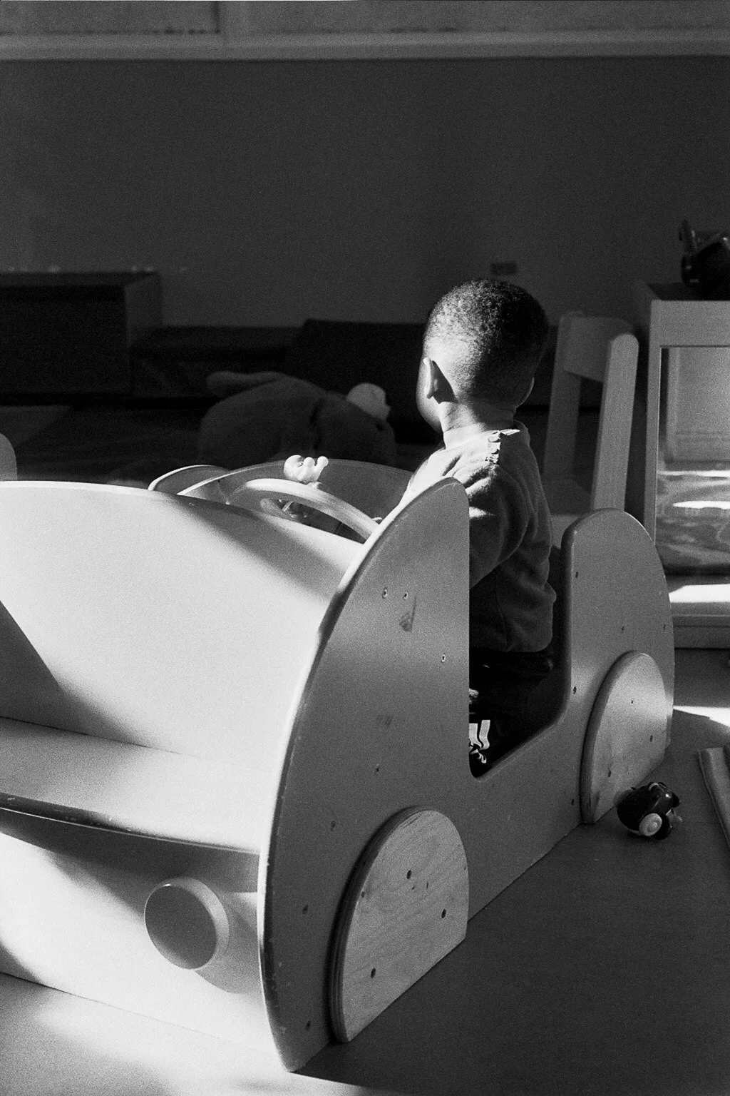 A young child sitting in a wooden toy car, looking away. Sunlight casts shadows in the room, with some furniture and toys visible.