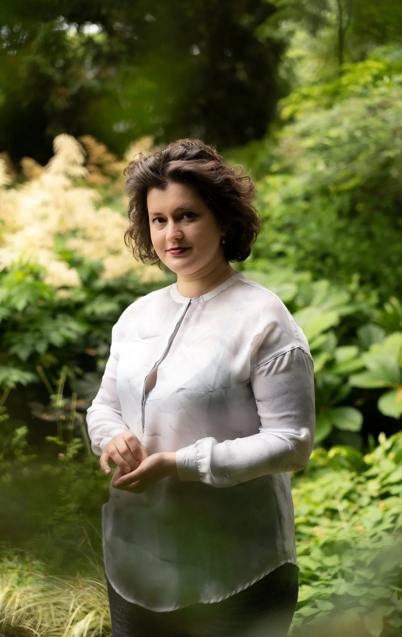 A woman with short, wavy brown hair standing outdoors in a lush, green garden or park, wearing a light-colored, long-sleeved blouse.