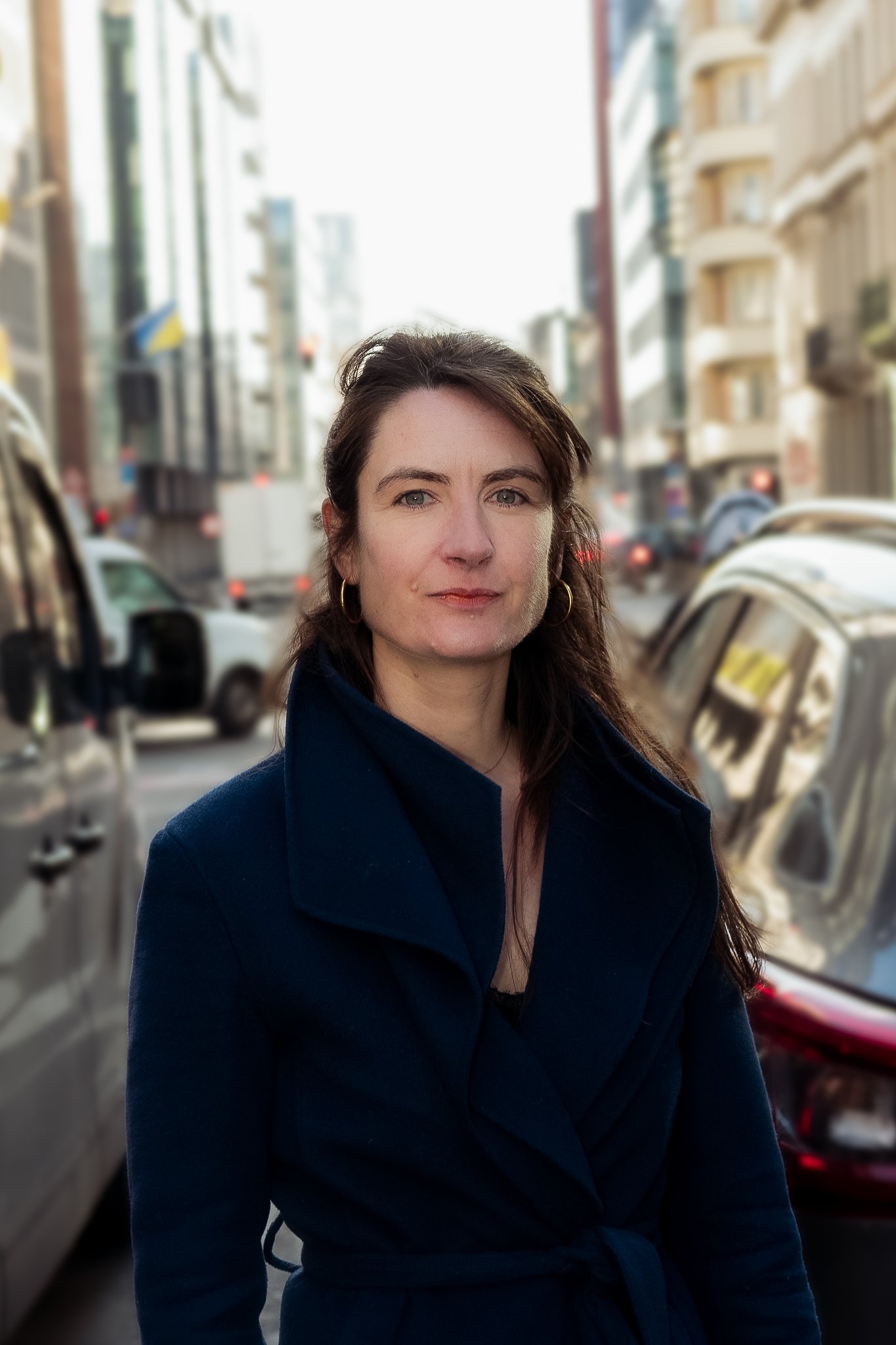 A woman with dark brown hair wearing a dark blue coat with a waist tie, standing on a city street with blurred cars and tall buildings in the background.