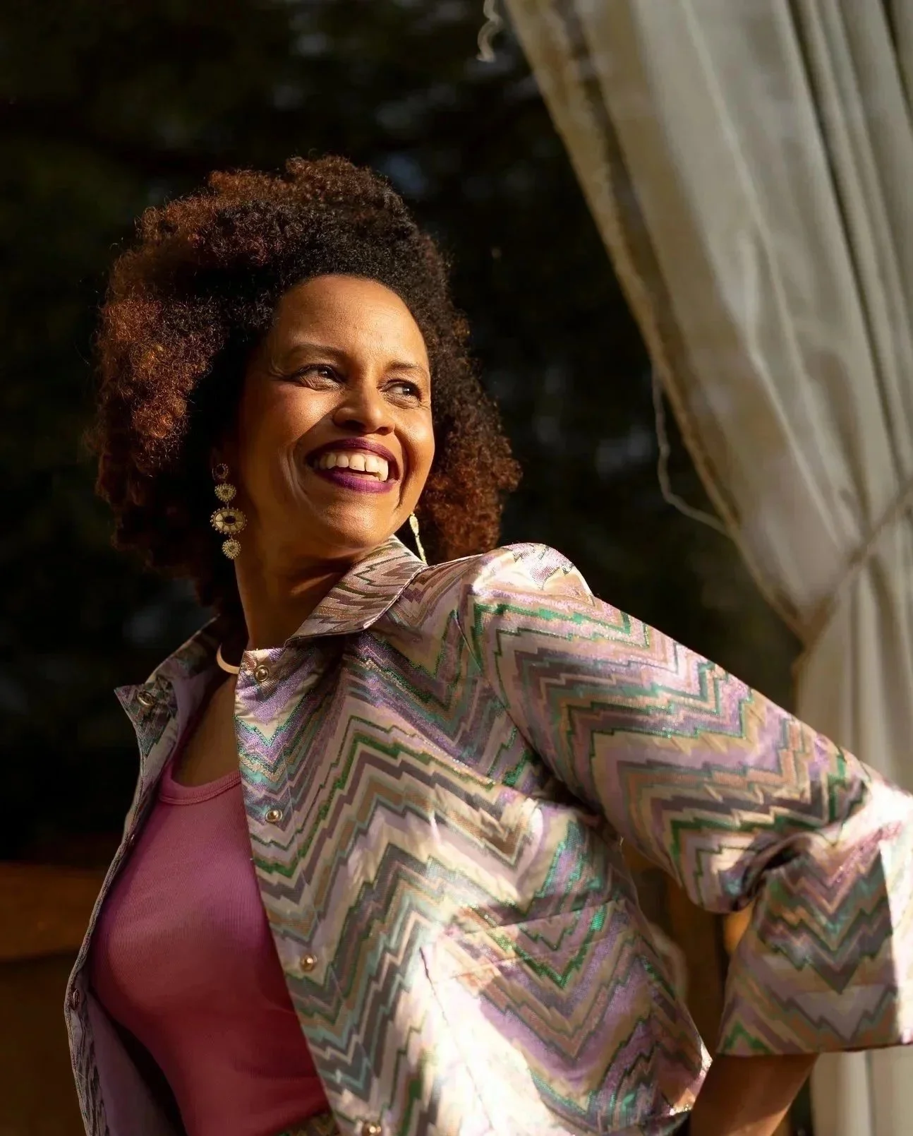 A woman with curly hair smiling and looking to the side, wearing a colorful patterned jacket and earrings, with a curtain in the background.