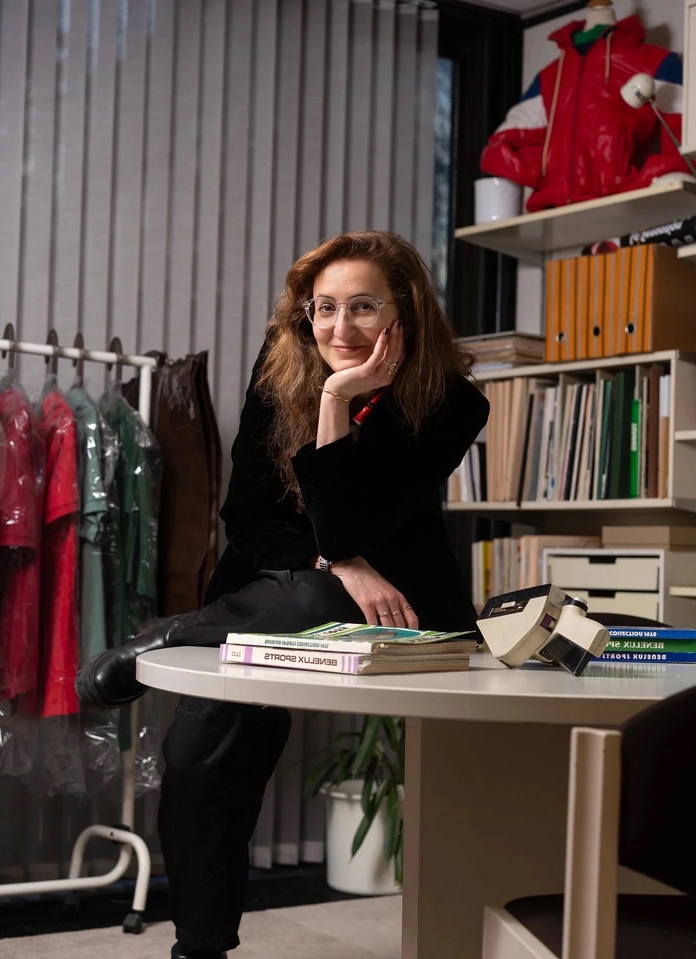 A woman with wavy brown hair, wearing glasses and a black outfit, sitting at a desk in a modern office or study space. She is smiling, resting her chin on her hand. On the desk are books, a telephone, and some papers. Behind her are shelves with books, folders, and a red jacket on a hanger, along with various office supplies.