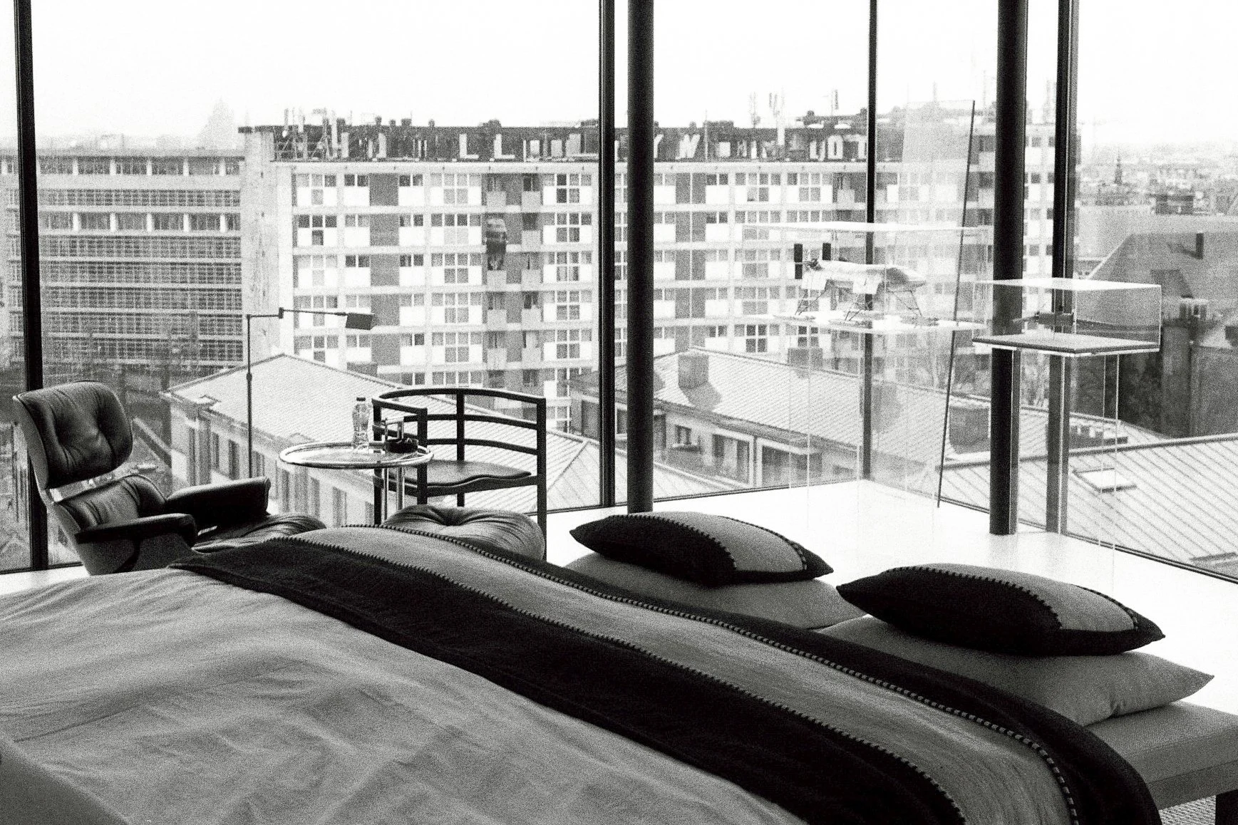 A modern bedroom with a large glass window overlooking city buildings. The bed has black and white bedding and pillows. There is a black leather chair, a small round table with a water bottle, and a balcony with a railing and some equipment outside.