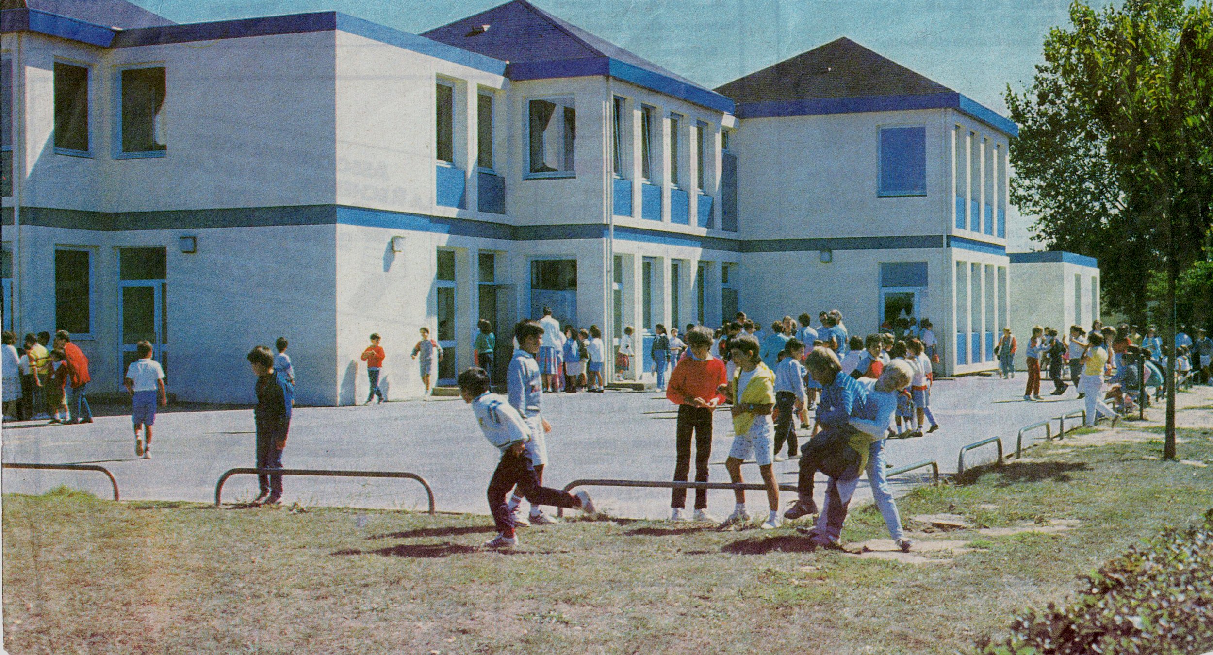 A school building with children outside, some walking and some waiting, on a sunny day. The school has white walls and blue trim, with a large group of students gathered near the entrance.