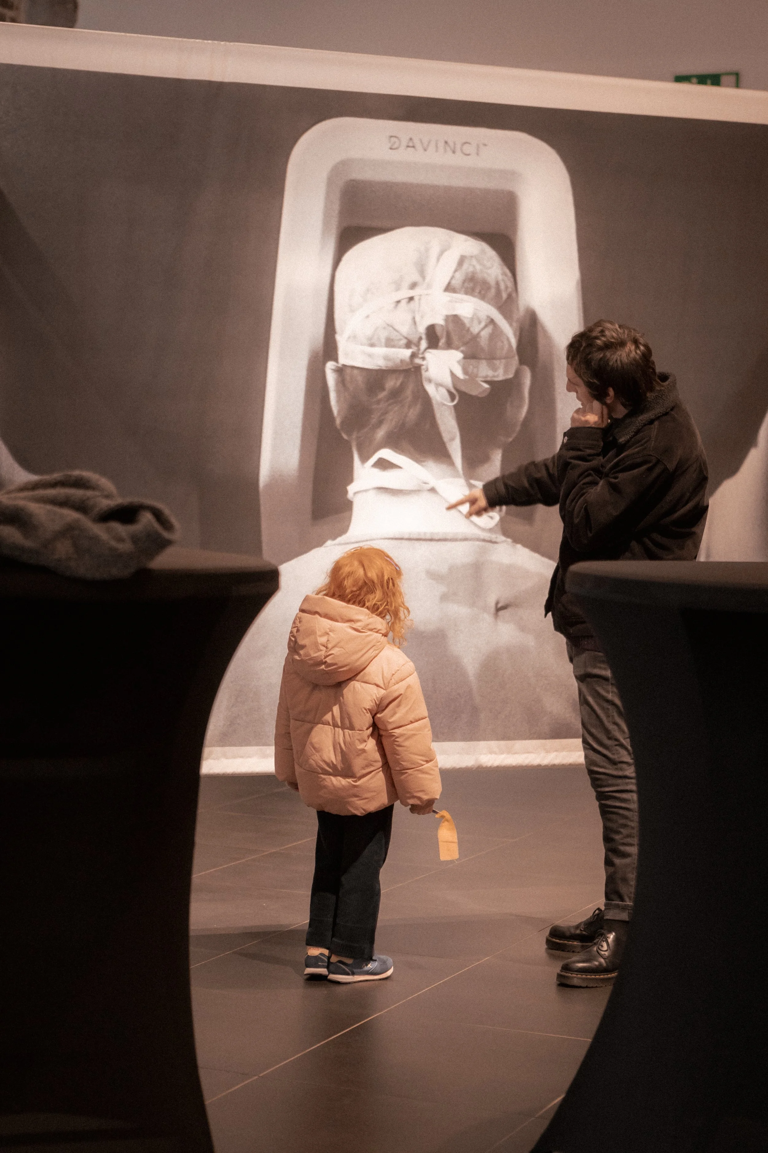 A woman and a young girl looking at an image of the back of a person wearing surgical scrubs and a cap in a museum or gallery setting.