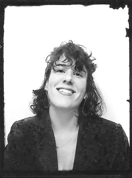 A woman with curly hair smiling, wearing a dark blazer, in a black and white photo with a rough border.