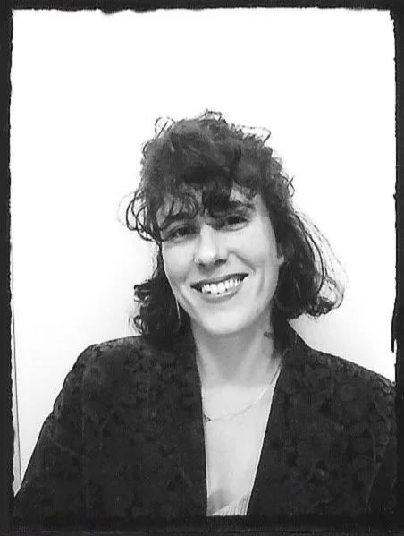 A woman with curly hair smiling in a black blazer, posed against a plain white background with a black border.