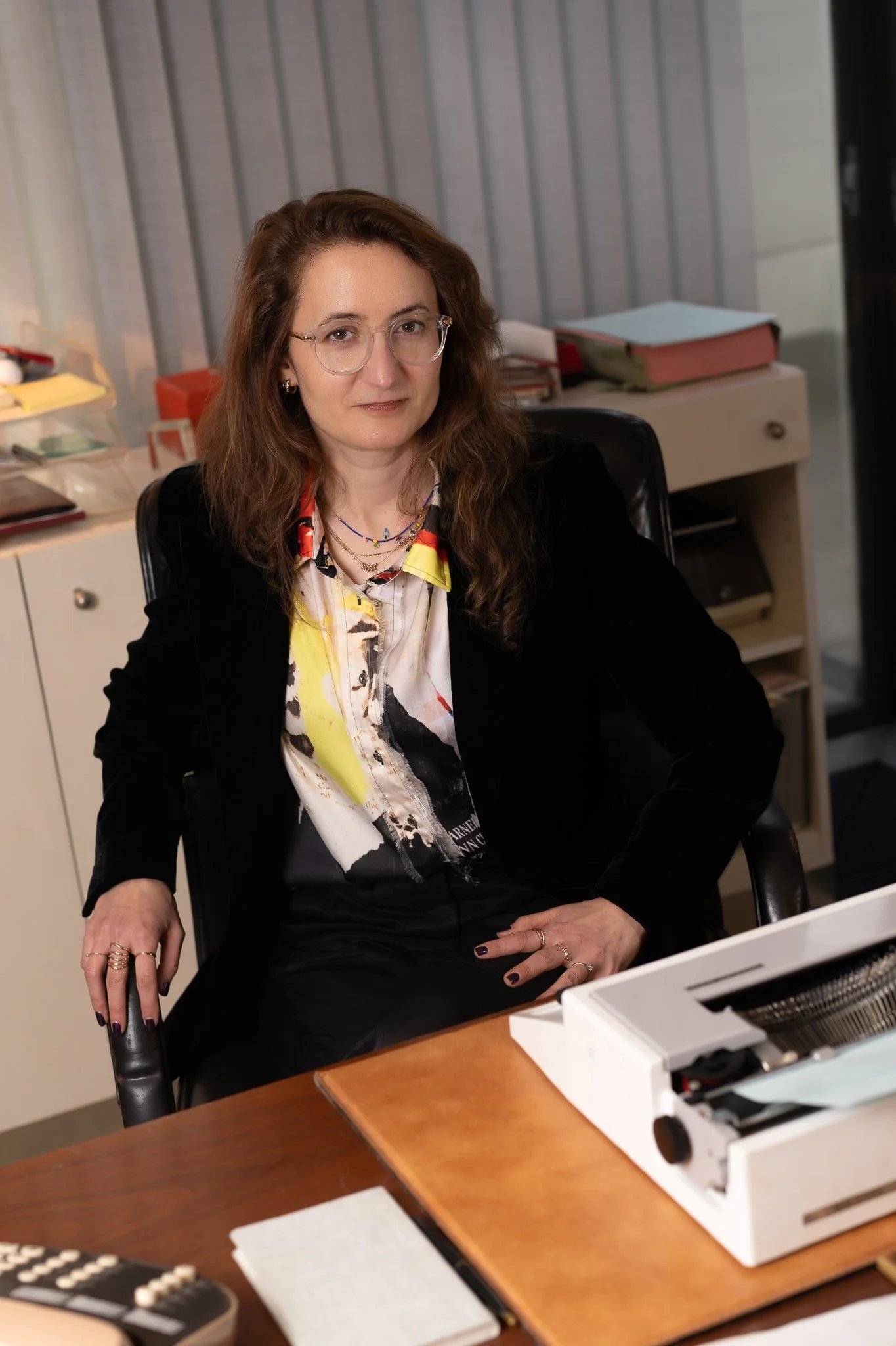 A woman with brown hair and glasses sitting at a desk with a typewriter, in an office space with files and papers behind her.
