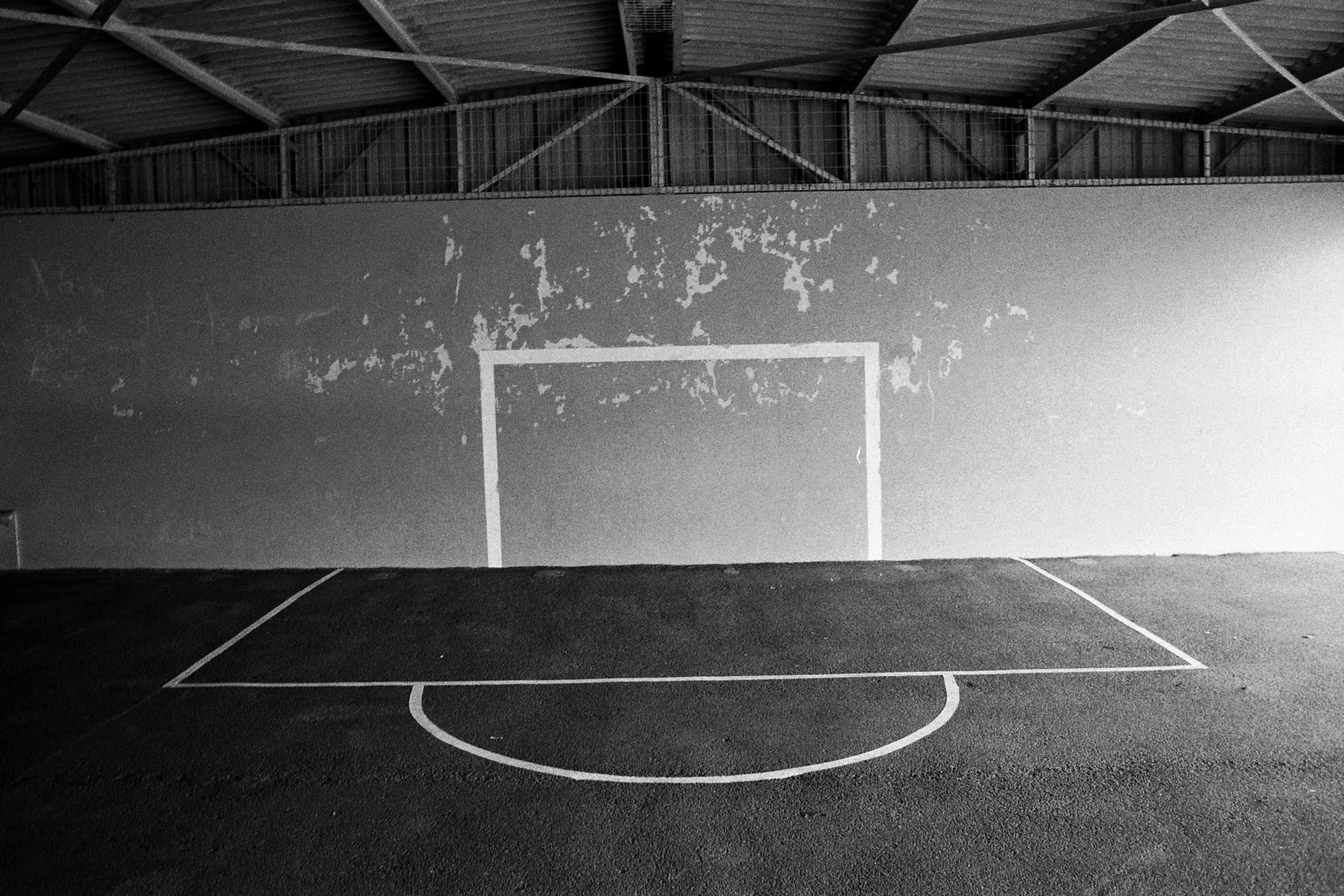 An indoor basketball court with a basketball hoop, painted lines on the floor, and a wall with peeling paint and graffiti reading 'A B X'.