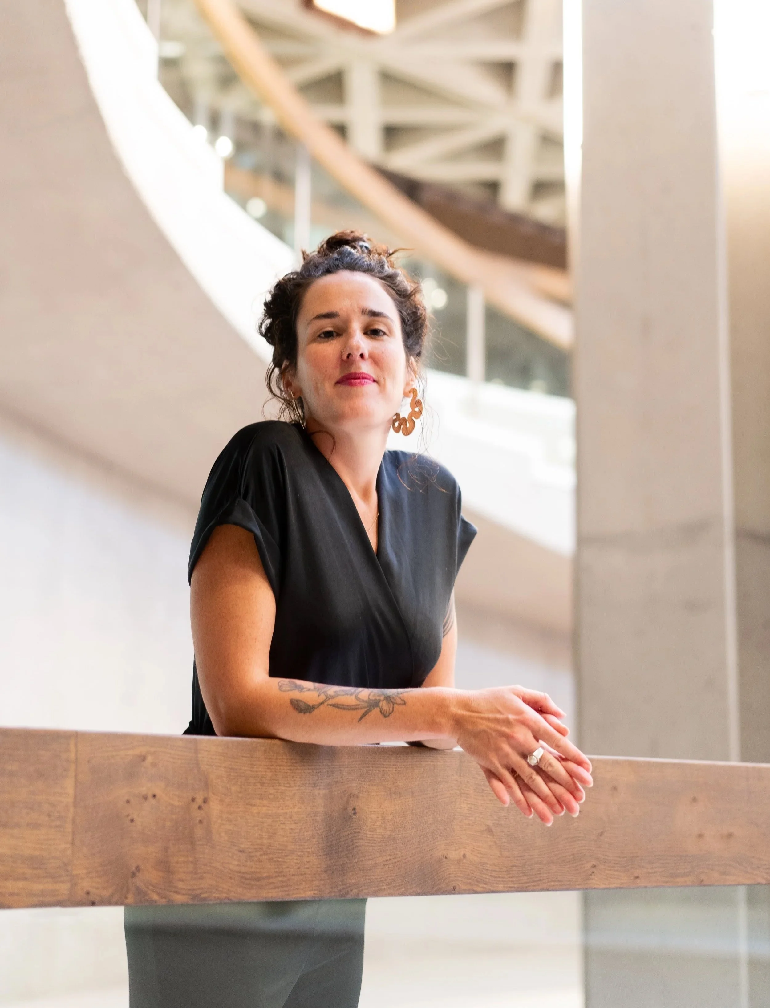 A woman with dark hair in an updo, wearing large earrings, a nose ring, and a black dress, leaning on a wooden bar or counter in an indoor setting with a modern architectural background.