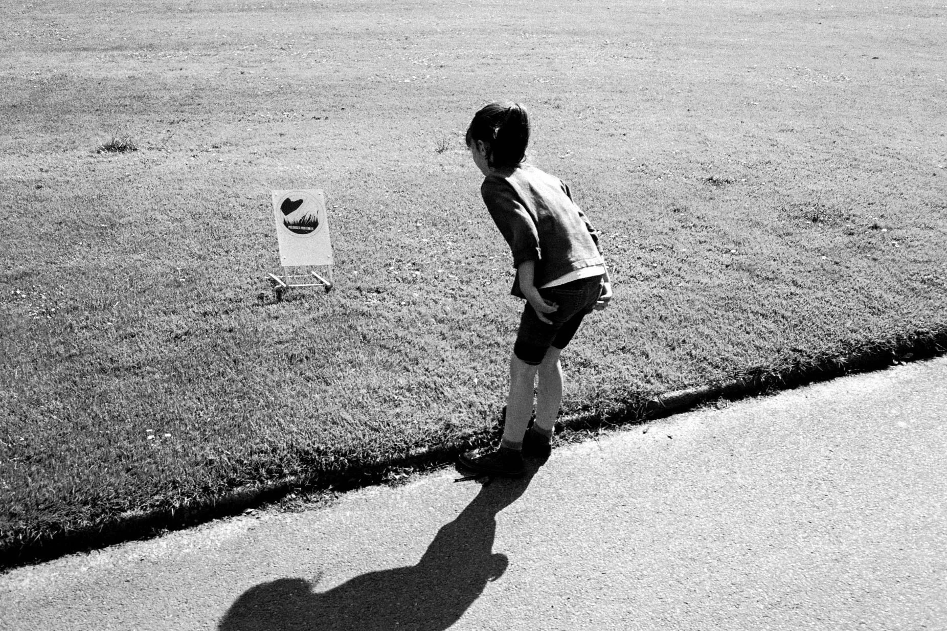 A boy in shorts and a jacket stands on a sidewalk, leaning over and looking down at a patch of grass. There is a lawn sign nearby with a drawn image and text, partially visible.