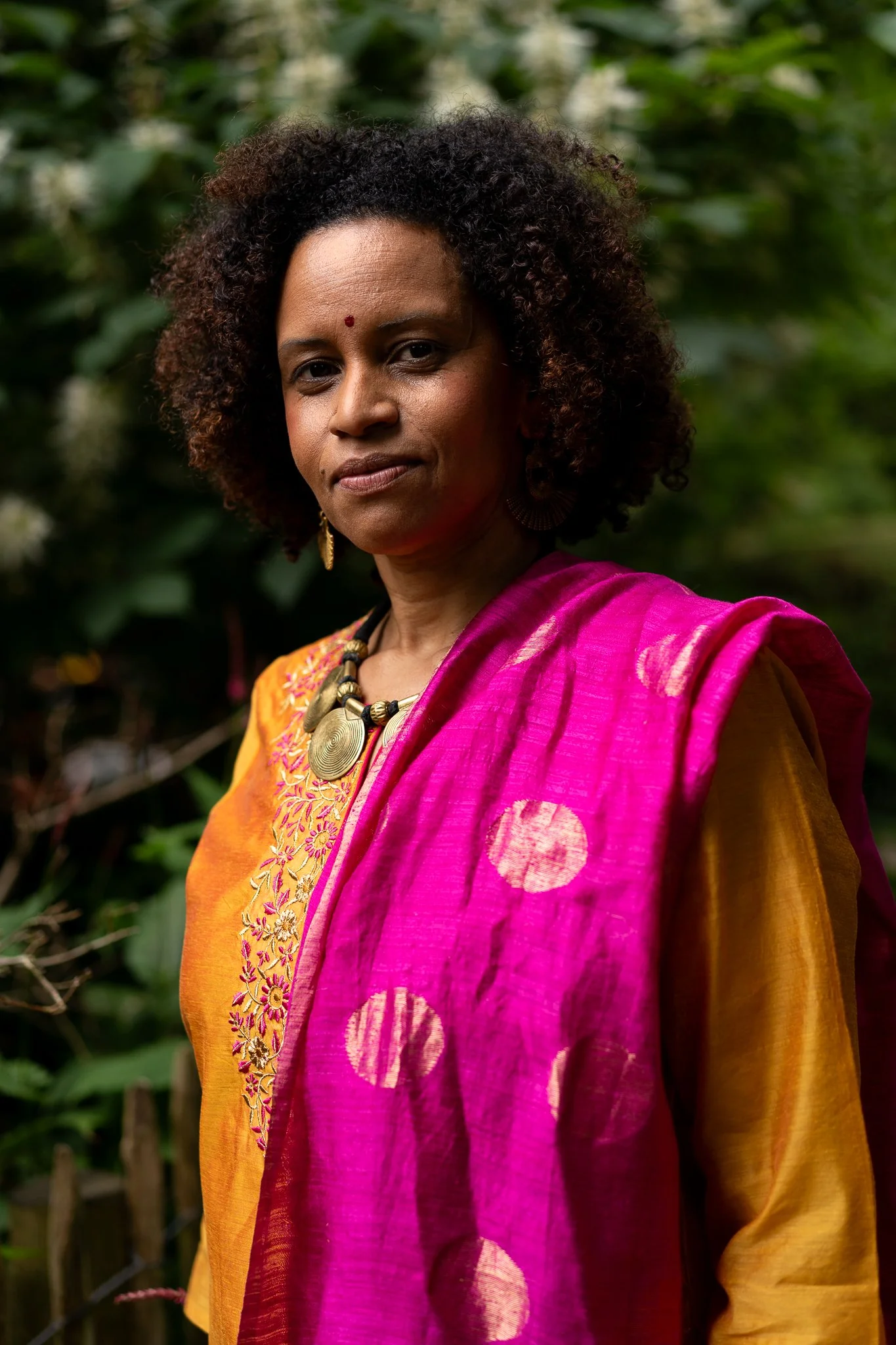 A woman with curly hair, wearing a traditional yellow and pink sari, standing outdoors with green foliage in the background.