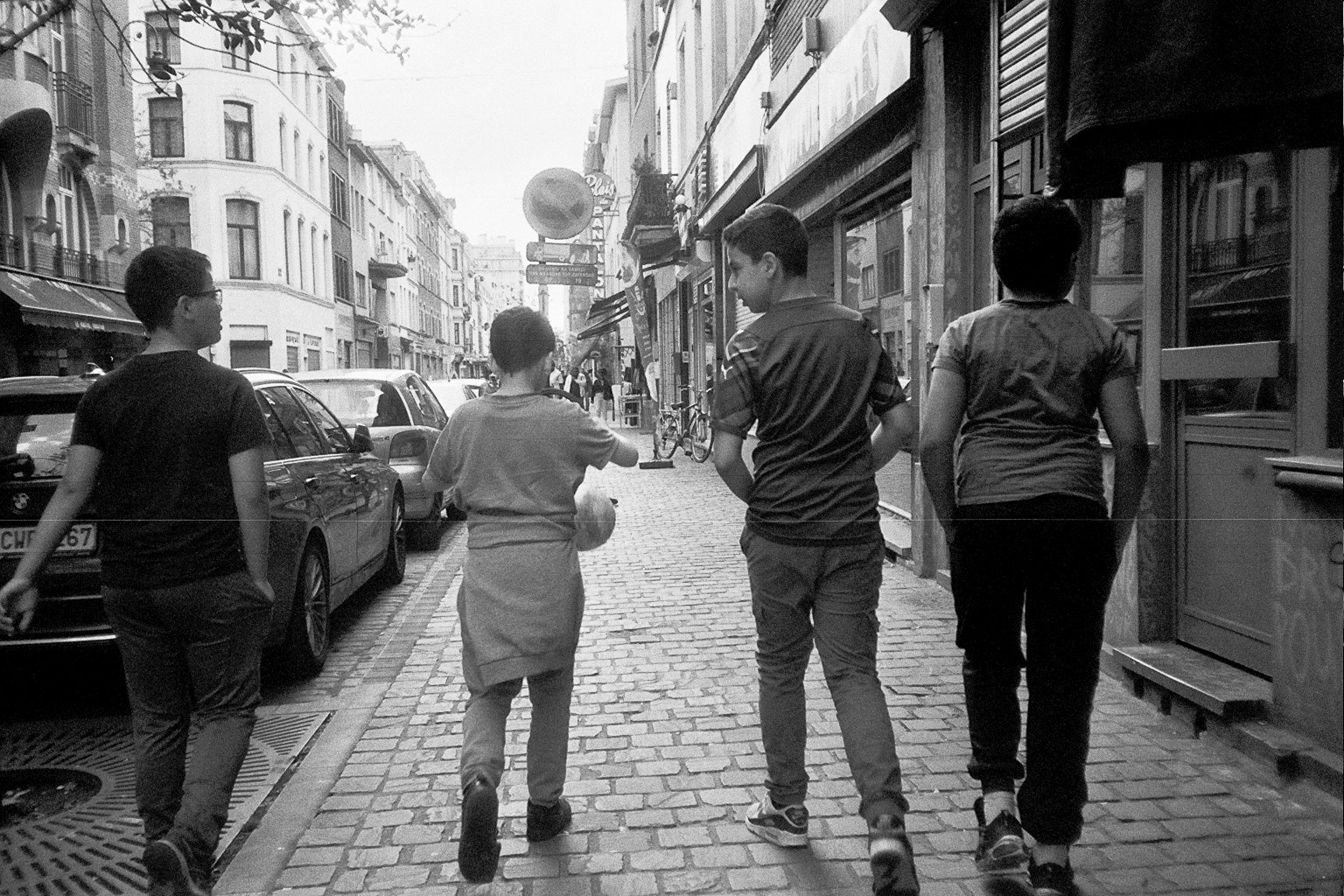 Four teenagers walk along a street in an urban area with cars parked on the side. The street is lined with buildings, shops, and signs, some with awnings.