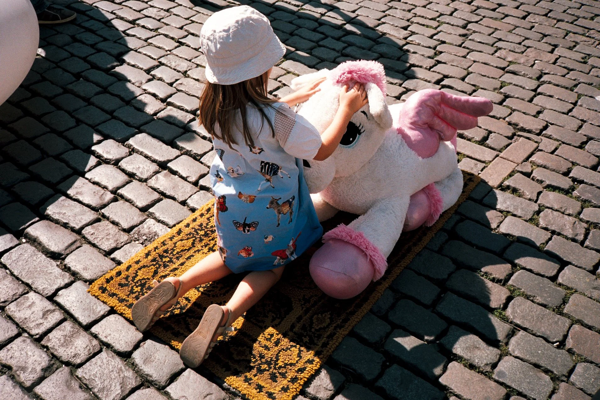 Child in a white dress with butterfly patterns, wearing a white sun hat, is kneeling on a yellow and black doormat on cobblestone street, hugging a large plush white unicorn with pink accents and big eyes.