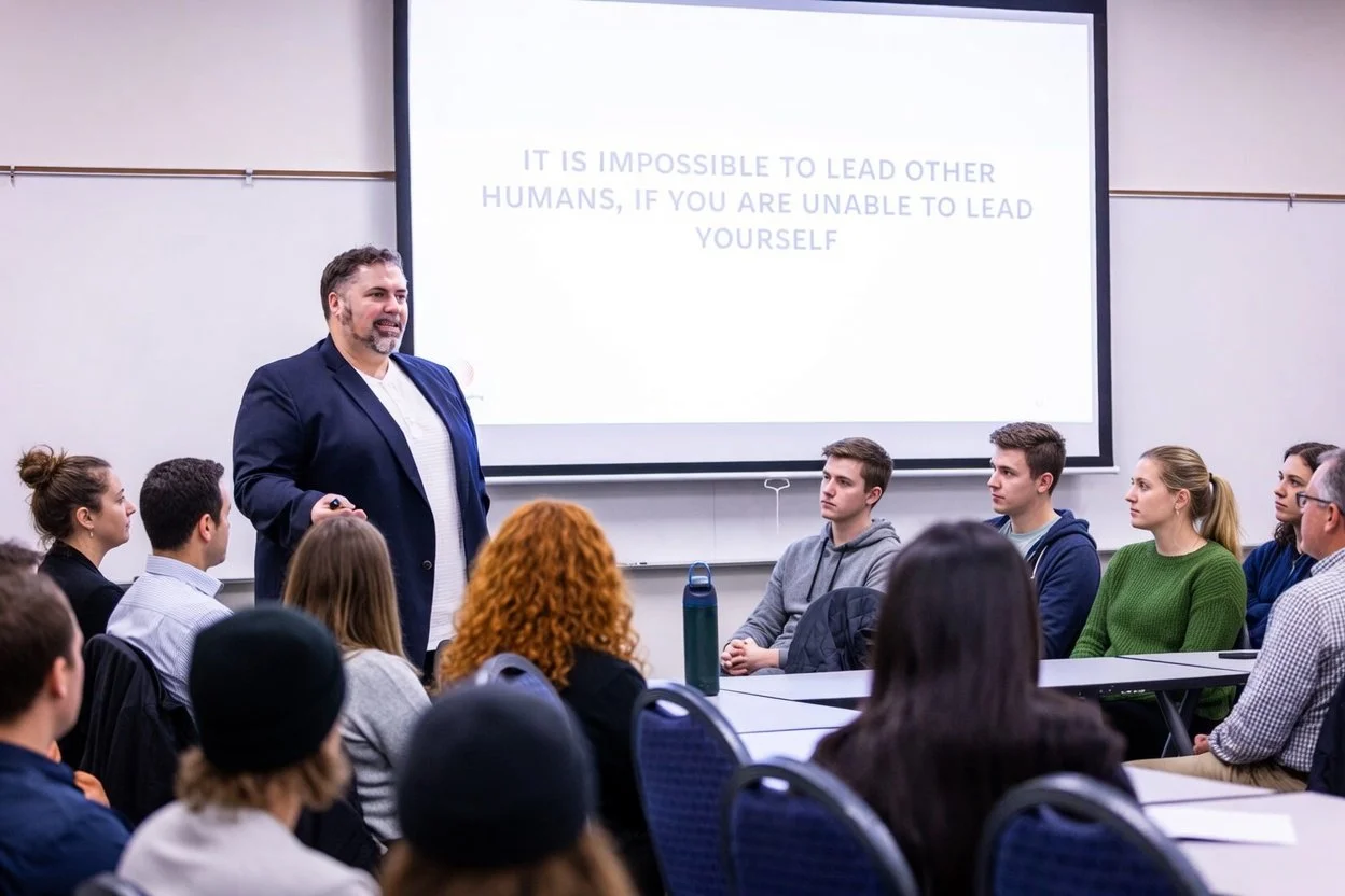 A man giving a presentation to a group of students in a classroom with a large screen displaying a quote about leadership.