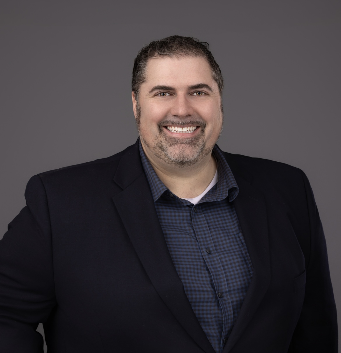 Portrait of a smiling man in a dark blazer and checkered shirt against a gray background.