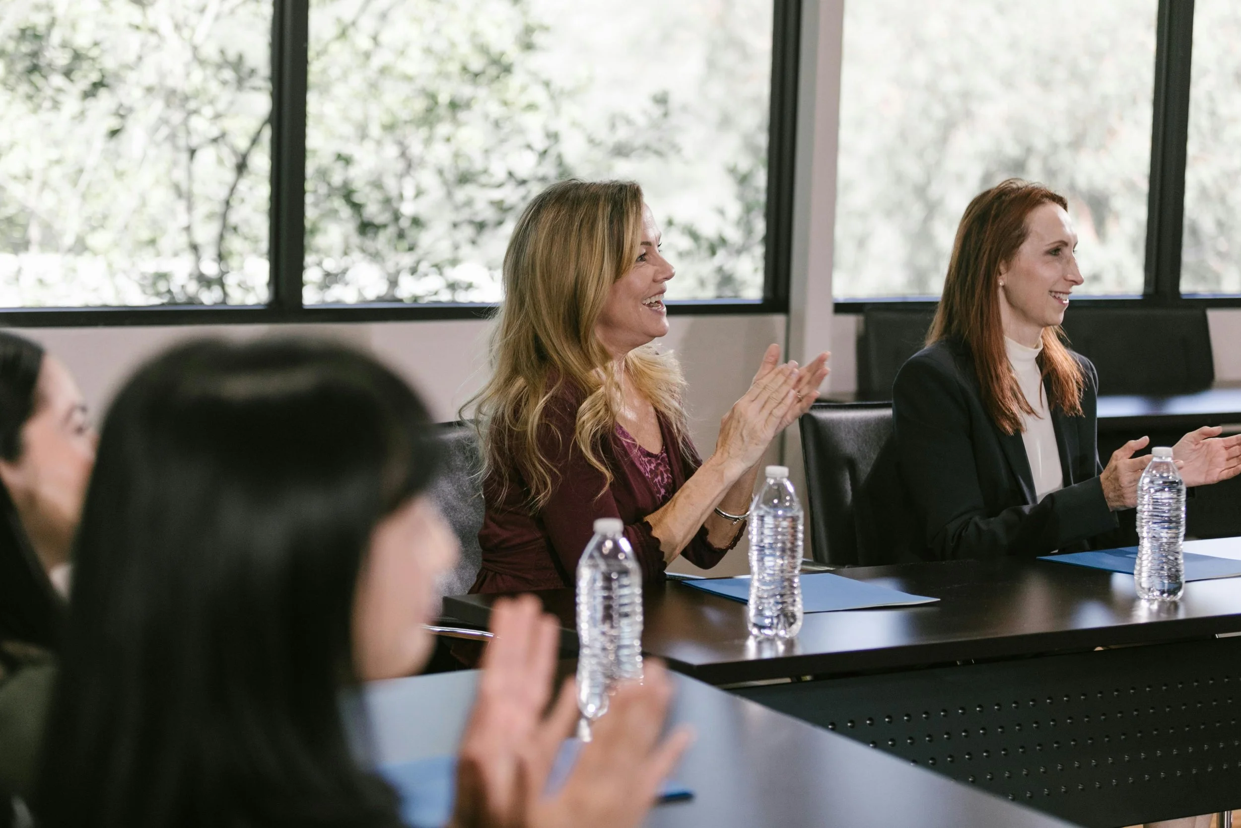 Group of women sitting at a conference table in a meeting room, clapping and smiling, with water bottles and blue folders in front of them.