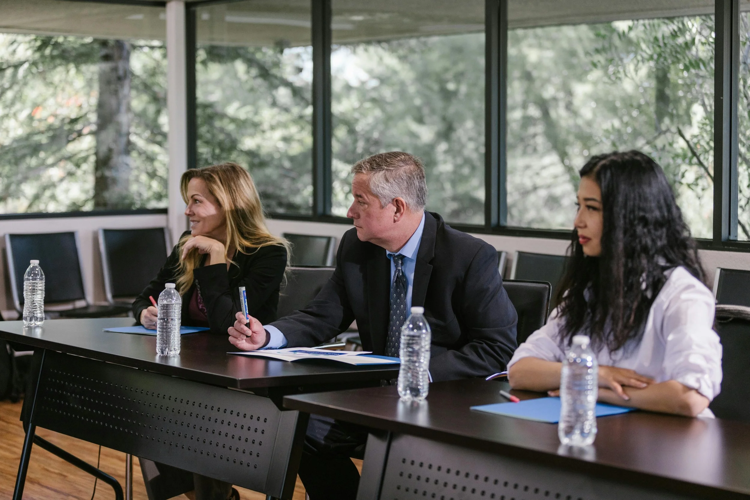 Three people sitting at a conference table with bottled water and blue folders, in a room with large windows overlooking a green outdoor scene.