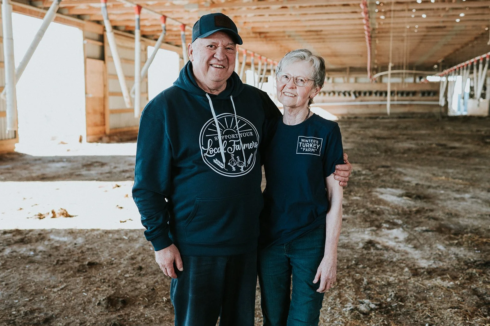 An elderly man and woman standing together inside a barn, smiling at the camera. The man is wearing a dark hoodie and a cap, while the woman is wearing glasses and a navy T-shirt. The barn has a wooden ceiling and a dirt floor.