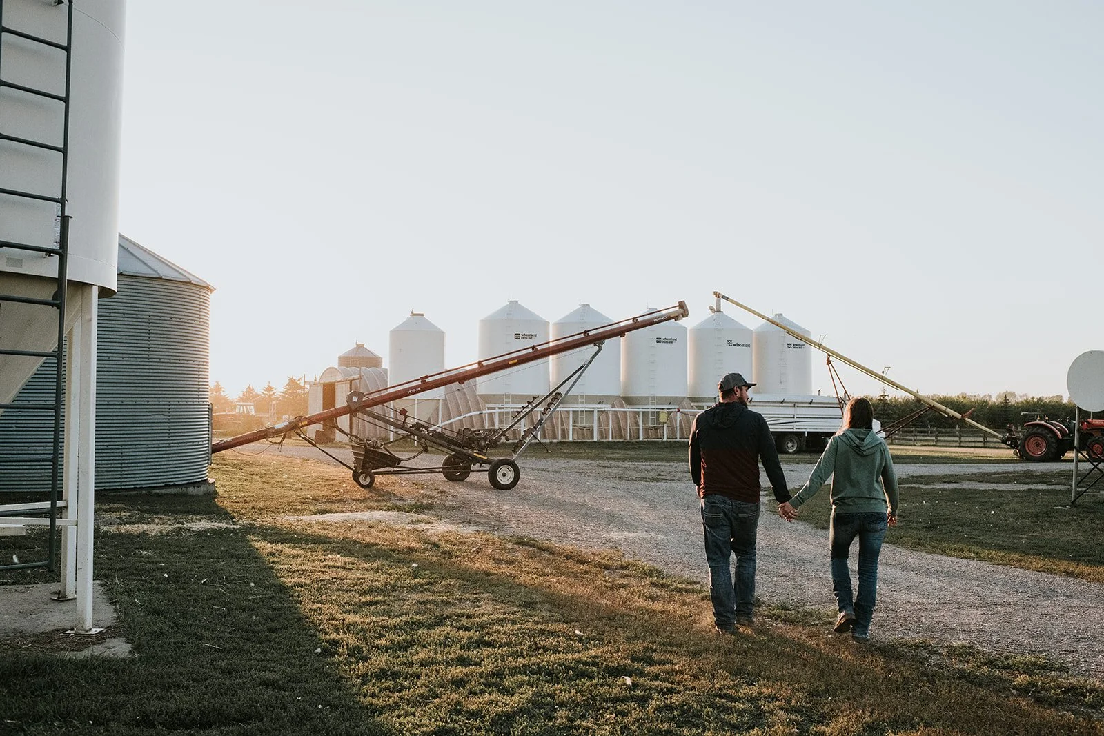 A couple holds hands and walks on a farm at sunset, with grain silos, farm equipment, and a tractor in the background.