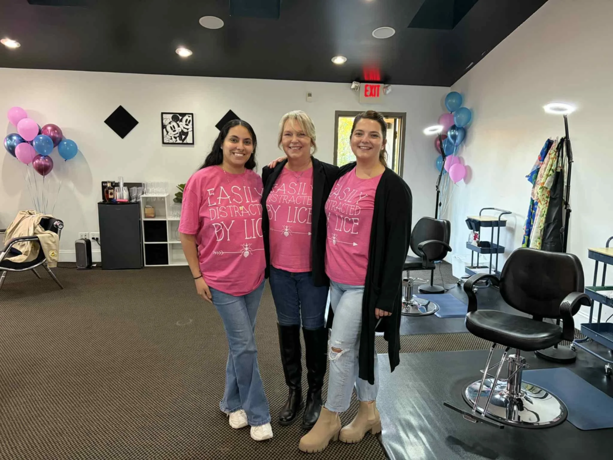 Three women wearing pink shirts that say 'EASILY DISTRACTED BY LICE,' standing together indoors, smiling and posing for a photo. The room has balloons, chairs, and tables, indicating a celebration or event.