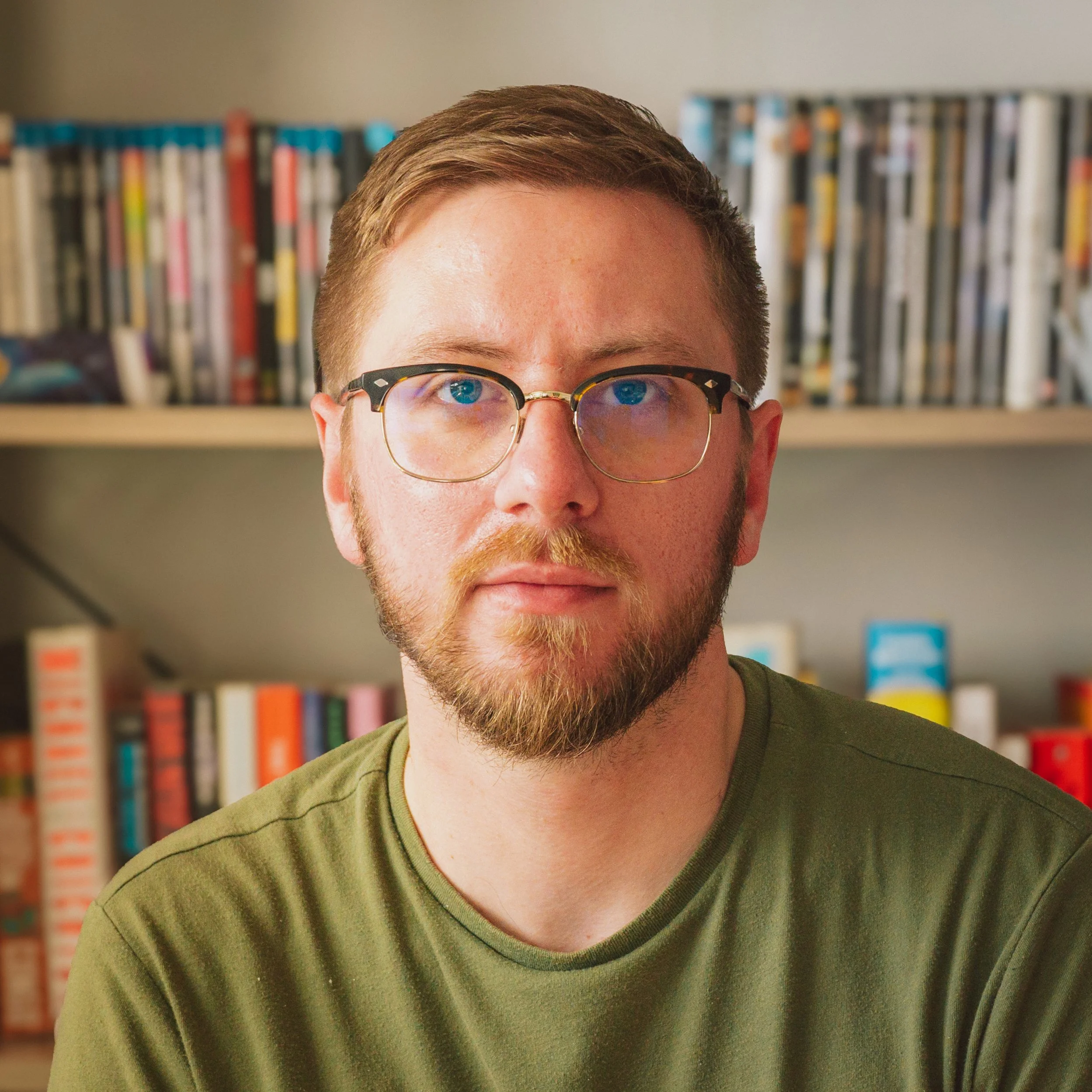 Close-up of a young man wearing glasses and a green shirt, with a bookshelf filled with books in the background.