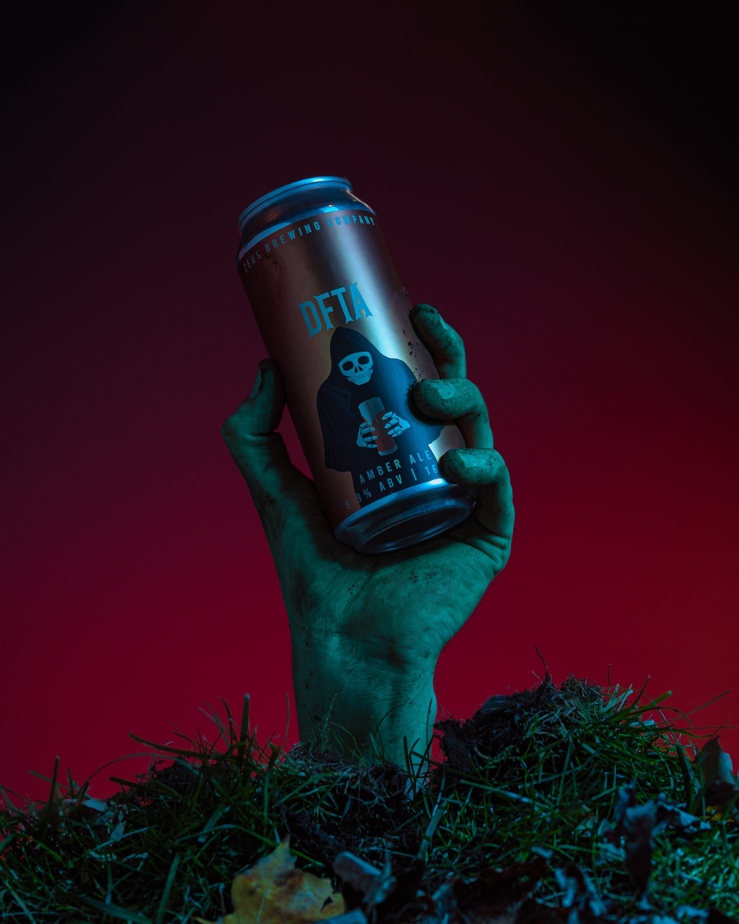 A hand with greenish fingers holding a can of Delta beer, with a dark sky background, illuminated by a bluish light, and grass and dirt at the bottom.