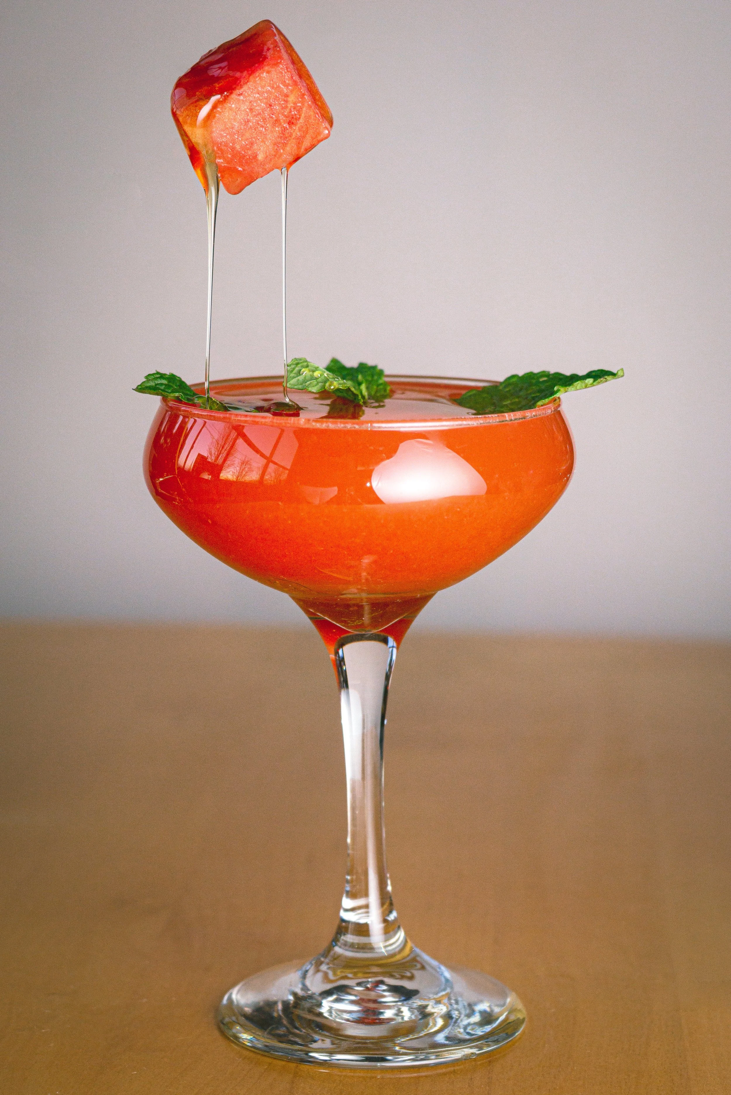 A pink cocktail in a coupe glass garnished with mint leaves, with a cube of watermelon being dipped into the drink.