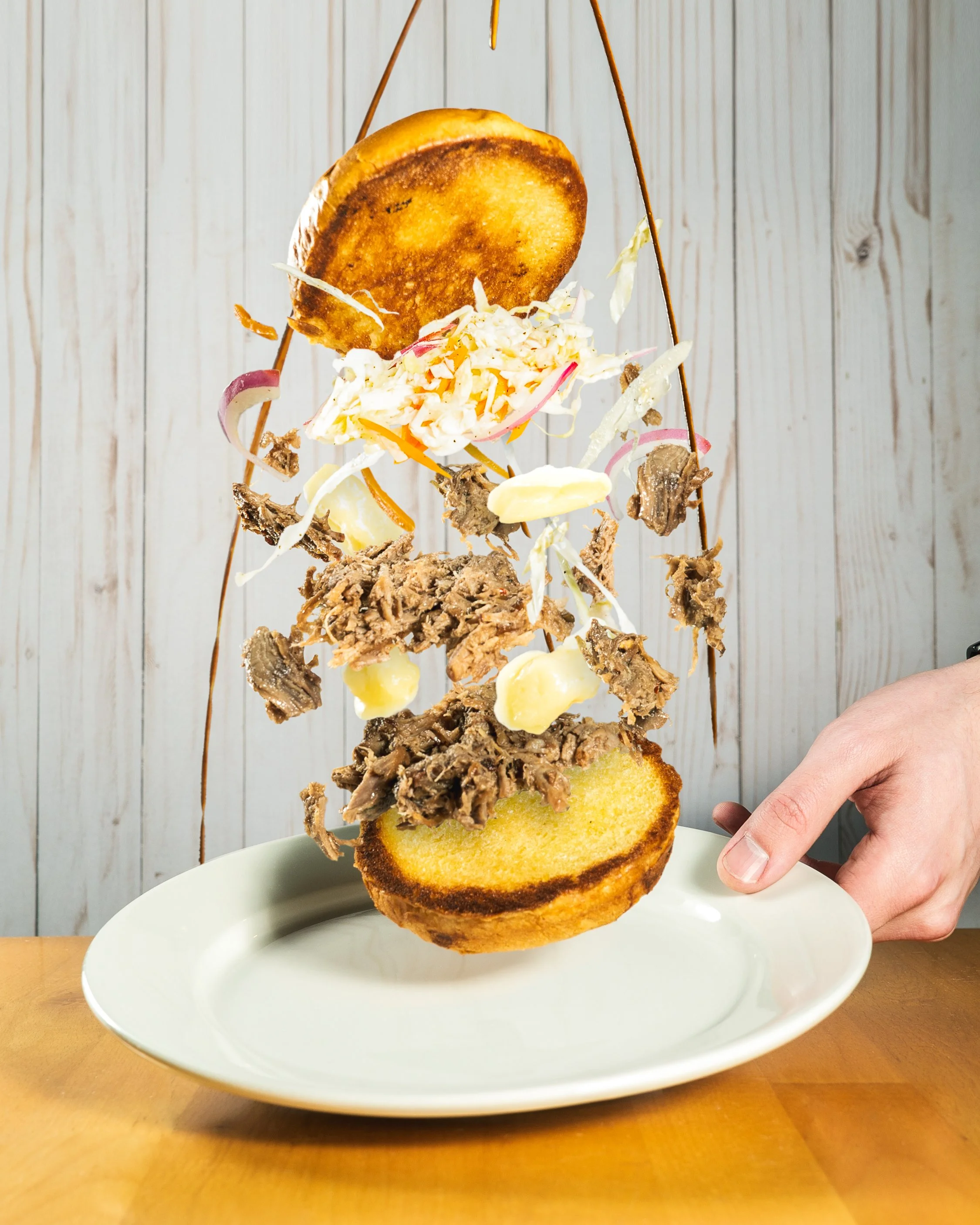 A person is balancing a burger vertically on a white plate, with a toasted bun at the bottom, layers of shredded cabbage and pulled meat in the middle, and a toasted bun on top, against a wooden wall background.