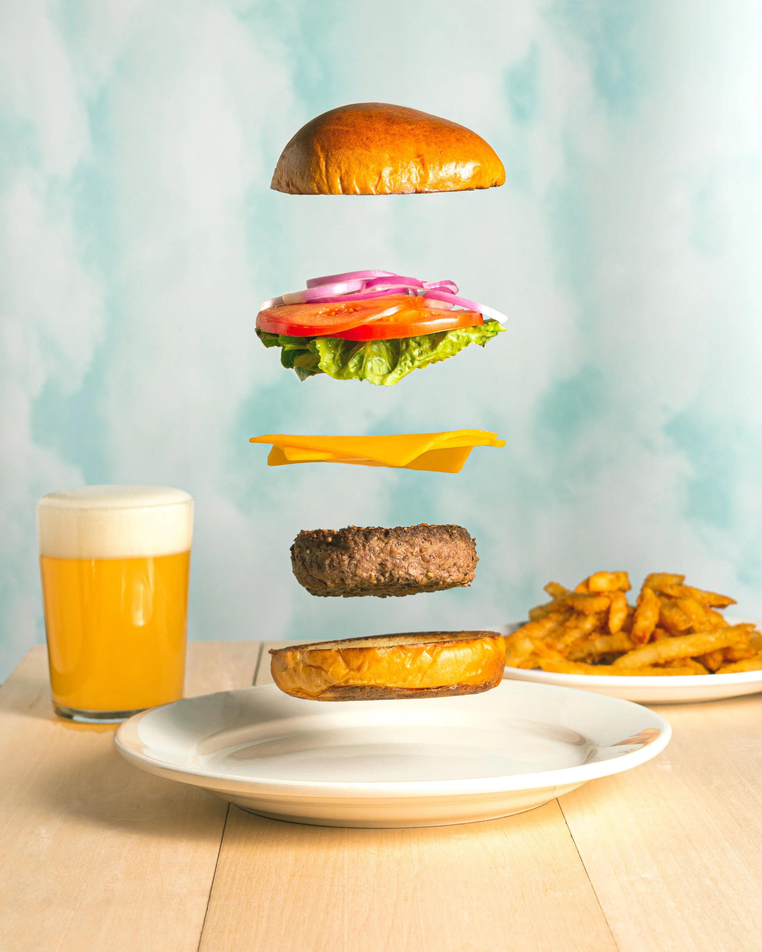 Floating burger ingredients including a top bun, tomato, onion, lettuce, hamburger patty, cheese, and bottom bun on a white plate, with a side of fries and a glass of beer in the background.