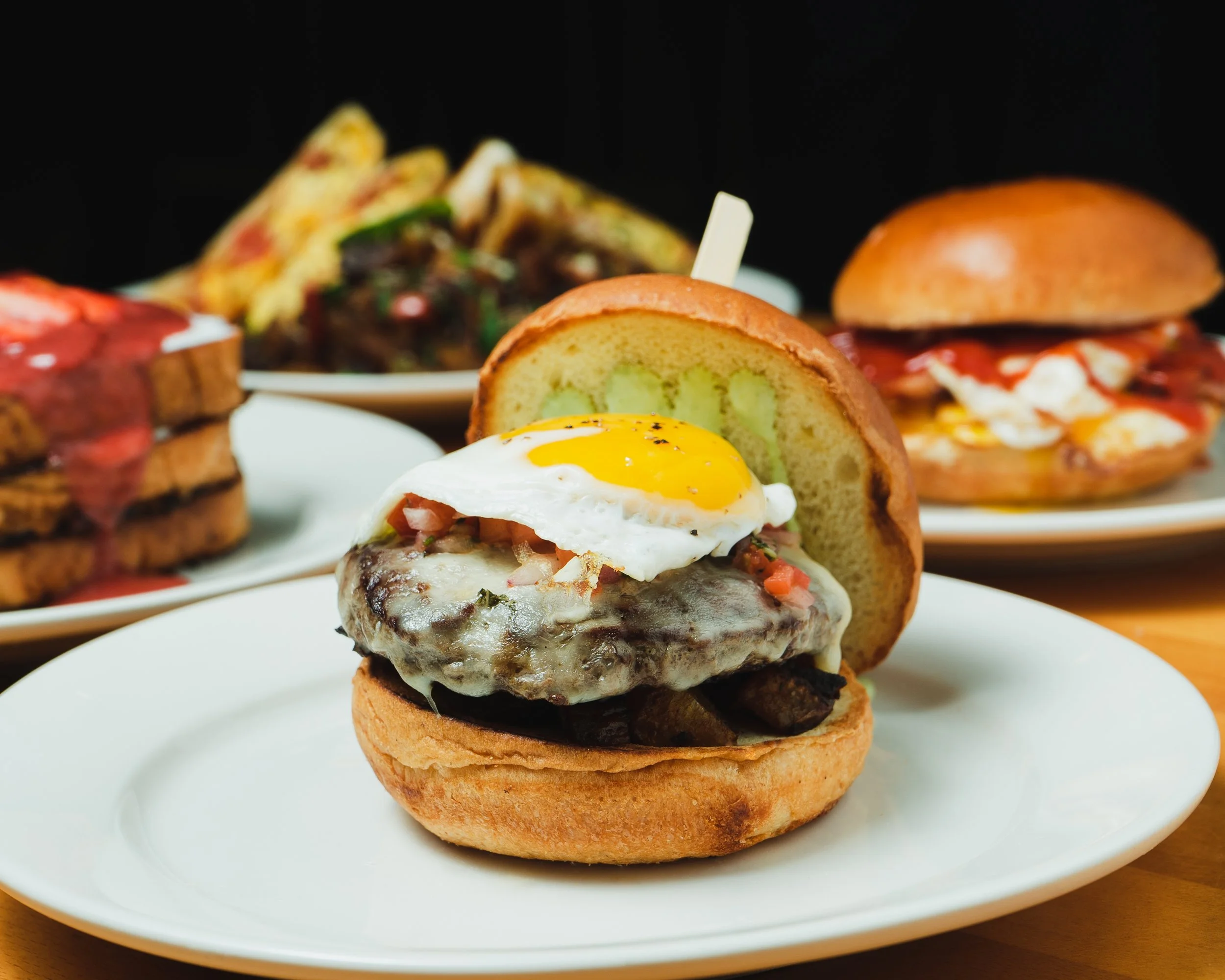 A close-up of a gourmet hamburger with a fried egg on top, served on a white plate, with other gourmet burgers in the background.