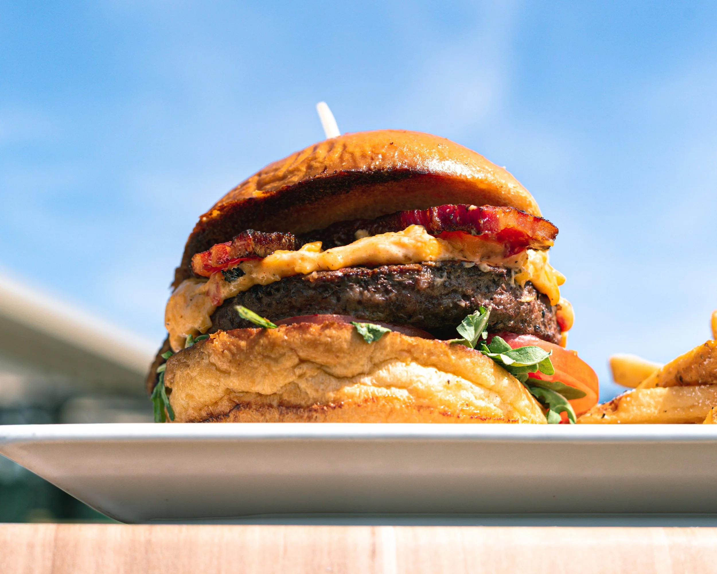 A close-up view of a tall burger with multiple beef patties, cheese, bacon, lettuce, and tomato, stacked high on a white plate against a clear blue sky background.