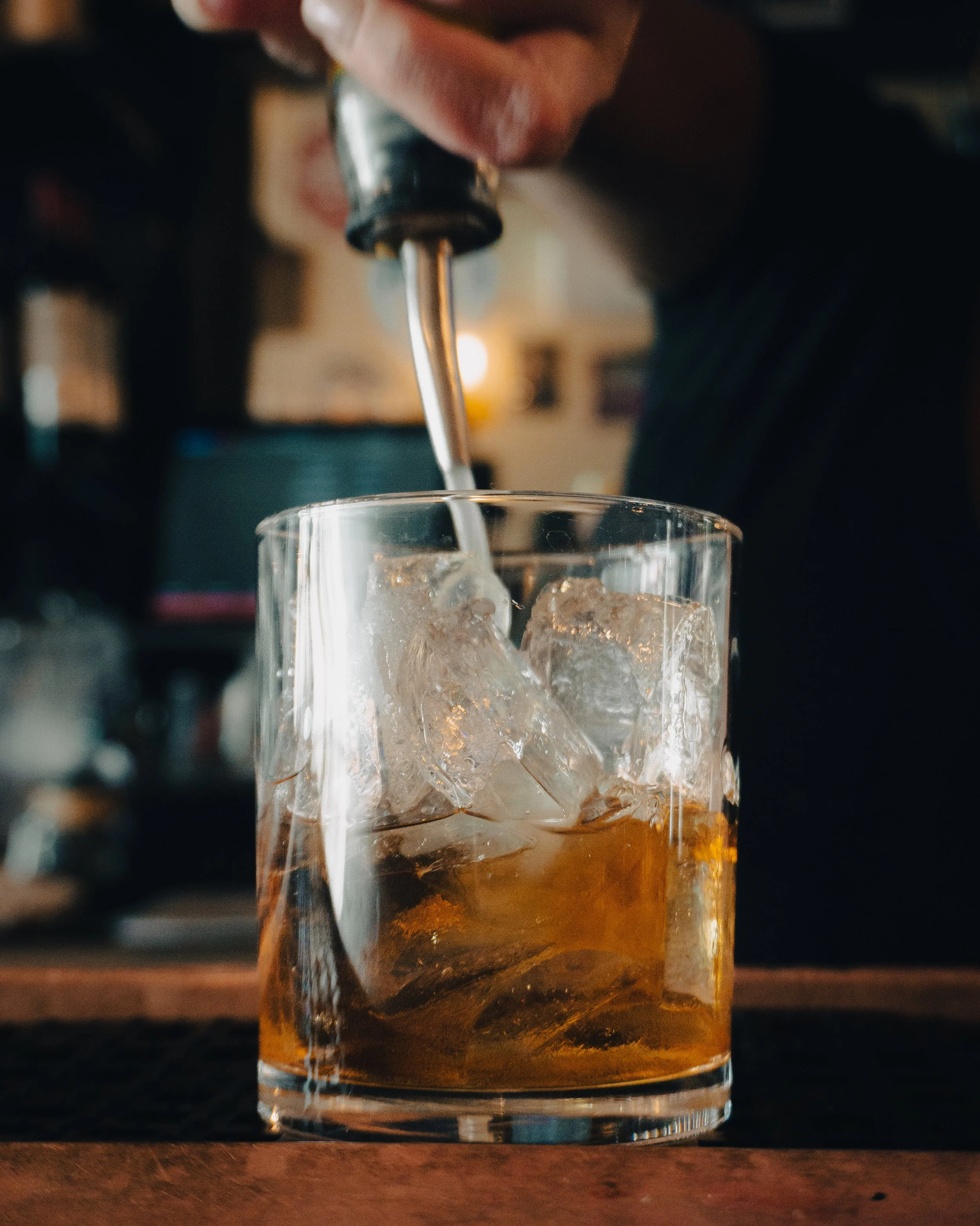 A glass of dark-colored beverage filled with ice cubes, with a hand pouring a clear liquid, possibly soda or water, into the glass.